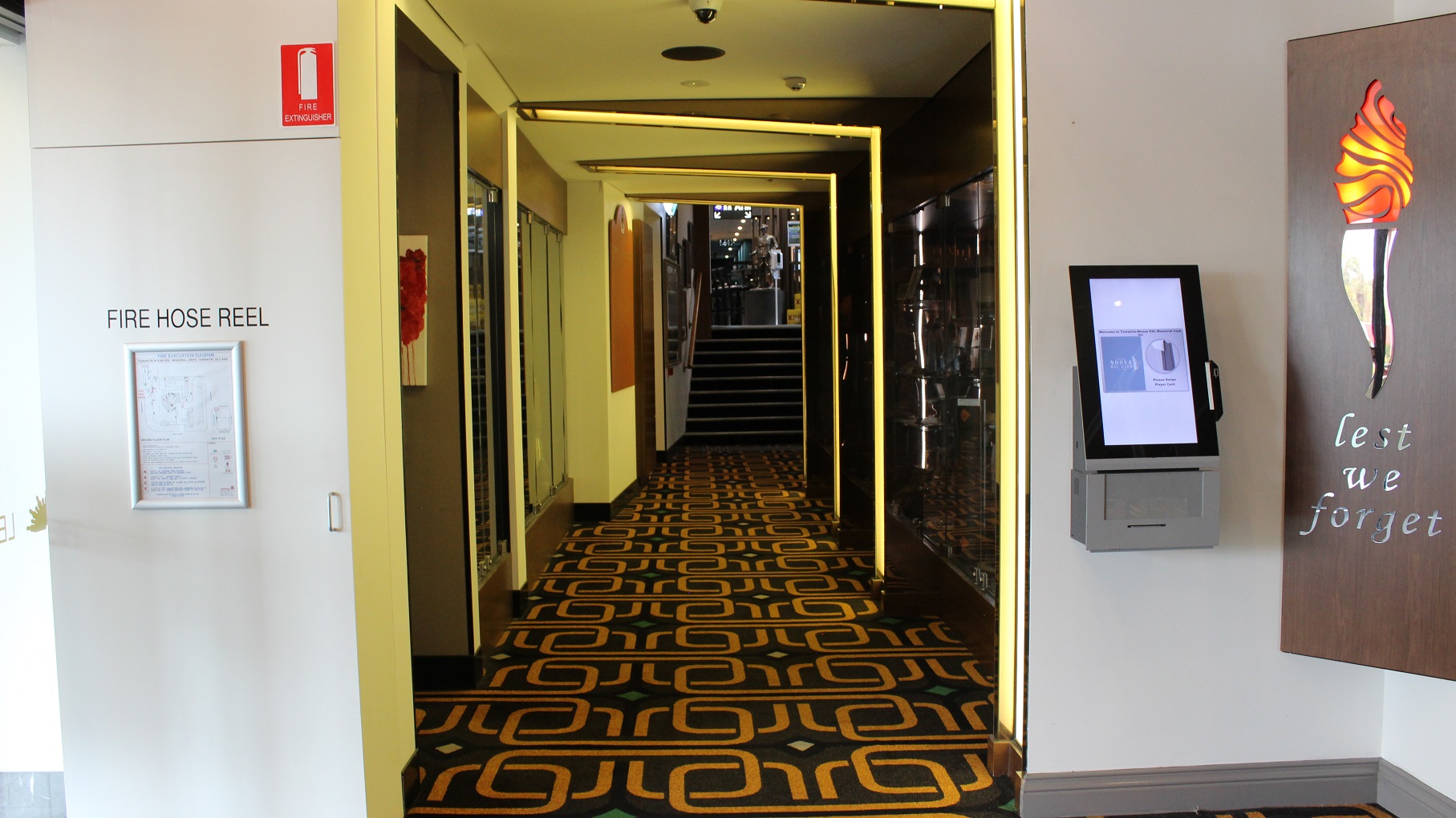 A lemon coloured-lit hallway leads to a grey-tiled step entryway with a check-in machine on the right. The artwork and glass patterns span both sides of the wall. A brown artwork, a fire hose reel, and an infographic are on the left wall. At the far end, is a sculpted soldier. The floor is covered in brown and gold. A lemon coloured-lit hallway leads to a grey-tiled step entryway with a check-in machine on the right. The artwork and glass patterns span both sides of the wall. A brown artwork, a fire hose reel, and an infographic are on the left wall. At the far end, is a sculpted soldier. The floor is covered in brown and gold.