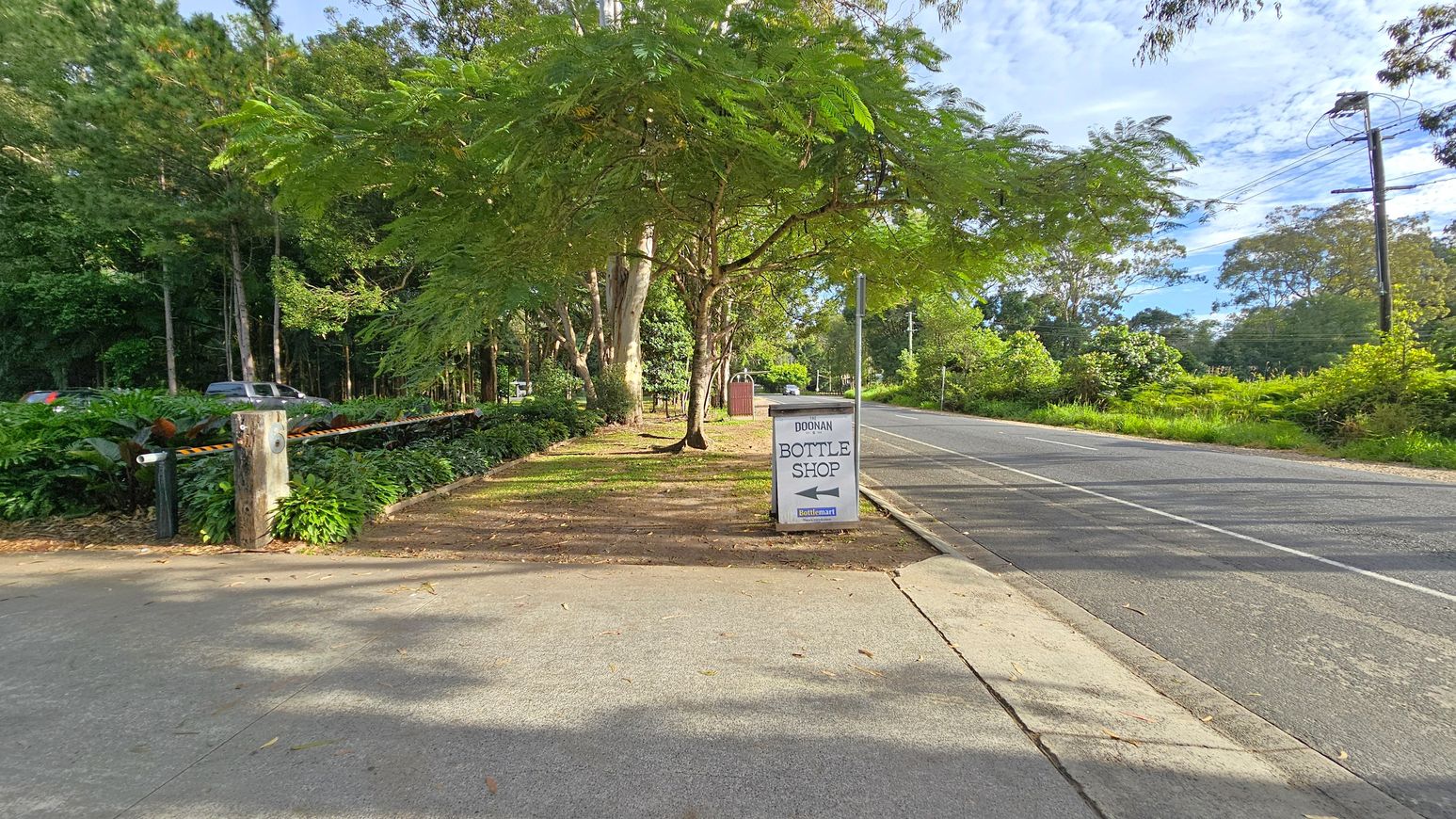 A road stretches into the distance on the right, bordered by lush greenery and trees. A sign on the pavement reads "Doonan Bottle Shop" with an arrow pointing left. On the left, a path runs parallel to the road, lined with dense foliage and a wooden post with a barrier. A vehicle is partially visible among the trees. The area is shaded by large trees, and the ground is covered with fallen leaves. A road stretches into the distance on the right, bordered by lush greenery and trees. A sign on the pavement reads "Doonan Bottle Shop" with an arrow pointing left. On the left, a path runs parallel to the road, lined with dense foliage and a wooden post with a barrier. A vehicle is partially visible among the trees. The area is shaded by large trees, and the ground is covered with fallen leaves.