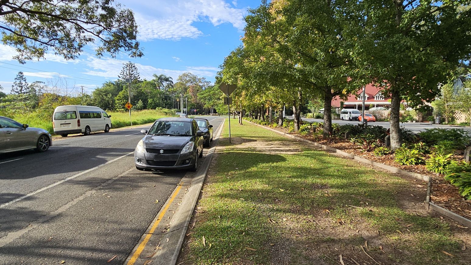 A street scene with a black car parked on the left side of the road. To the right, there's a grassy verge with trees lining the pavement. In the background, a white van and a silver car are driving on the road. Further back, a red-roofed building is partially visible behind more trees and shrubs. The road is bordered by a yellow line, and the area is well-shaded by the trees. A street scene with a black car parked on the left side of the road. To the right, there's a grassy verge with trees lining the pavement. In the background, a white van and a silver car are driving on the road. Further back, a red-roofed building is partially visible behind more trees and shrubs. The road is bordered by a yellow line, and the area is well-shaded by the trees.