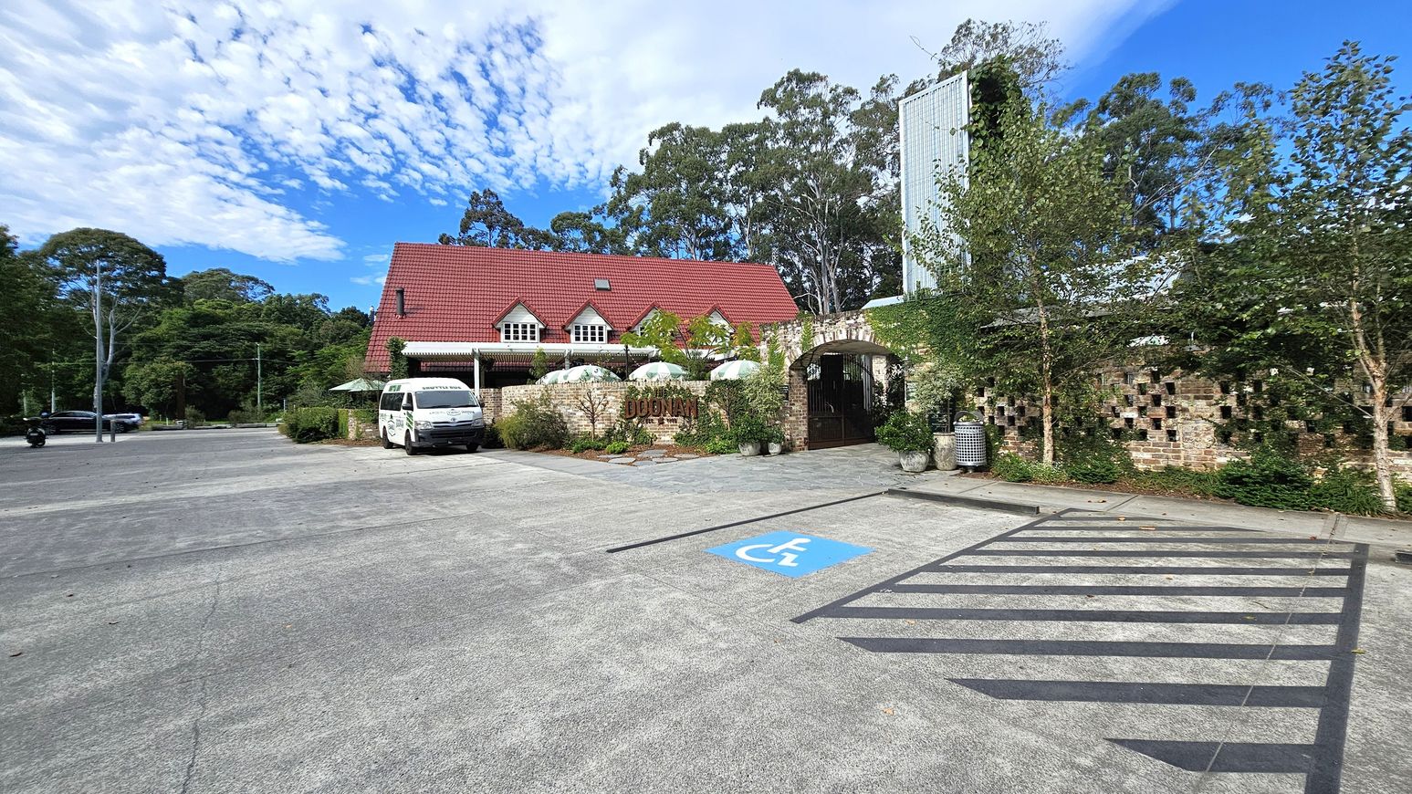A building with a red gabled roof and multiple dormer windows is in the centre. In front, a white van is parked near a brick wall with a sign. Several white umbrellas are visible behind the wall. On the right, there is a patterned brick wall with greenery and trees. A blue disabled parking space is marked on the concrete ground in the foreground. Tall trees are in the background, and a few vehicles are on the left side of the parking area. A building with a red gabled roof and multiple dormer windows is in the centre. In front, a white van is parked near a brick wall with a sign. Several white umbrellas are visible behind the wall. On the right, there is a patterned brick wall with greenery and trees. A blue disabled parking space is marked on the concrete ground in the foreground. Tall trees are in the background, and a few vehicles are on the left side of the parking area.