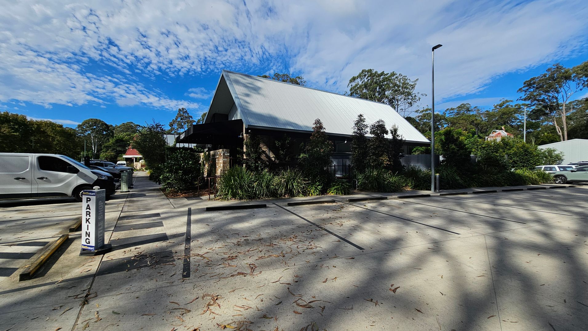 A parking area with a sign marked "PARKING" on the left. Several vehicles, including a white van, are parked. In the centre, a building with a steep, corrugated metal roof is surrounded by greenery. The right side shows more parking spaces and a tall streetlamp. The ground is concrete with scattered leaves and shadows from nearby trees. A parking area with a sign marked "PARKING" on the left. Several vehicles, including a white van, are parked. In the centre, a building with a steep, corrugated metal roof is surrounded by greenery. The right side shows more parking spaces and a tall streetlamp. The ground is concrete with scattered leaves and shadows from nearby trees.