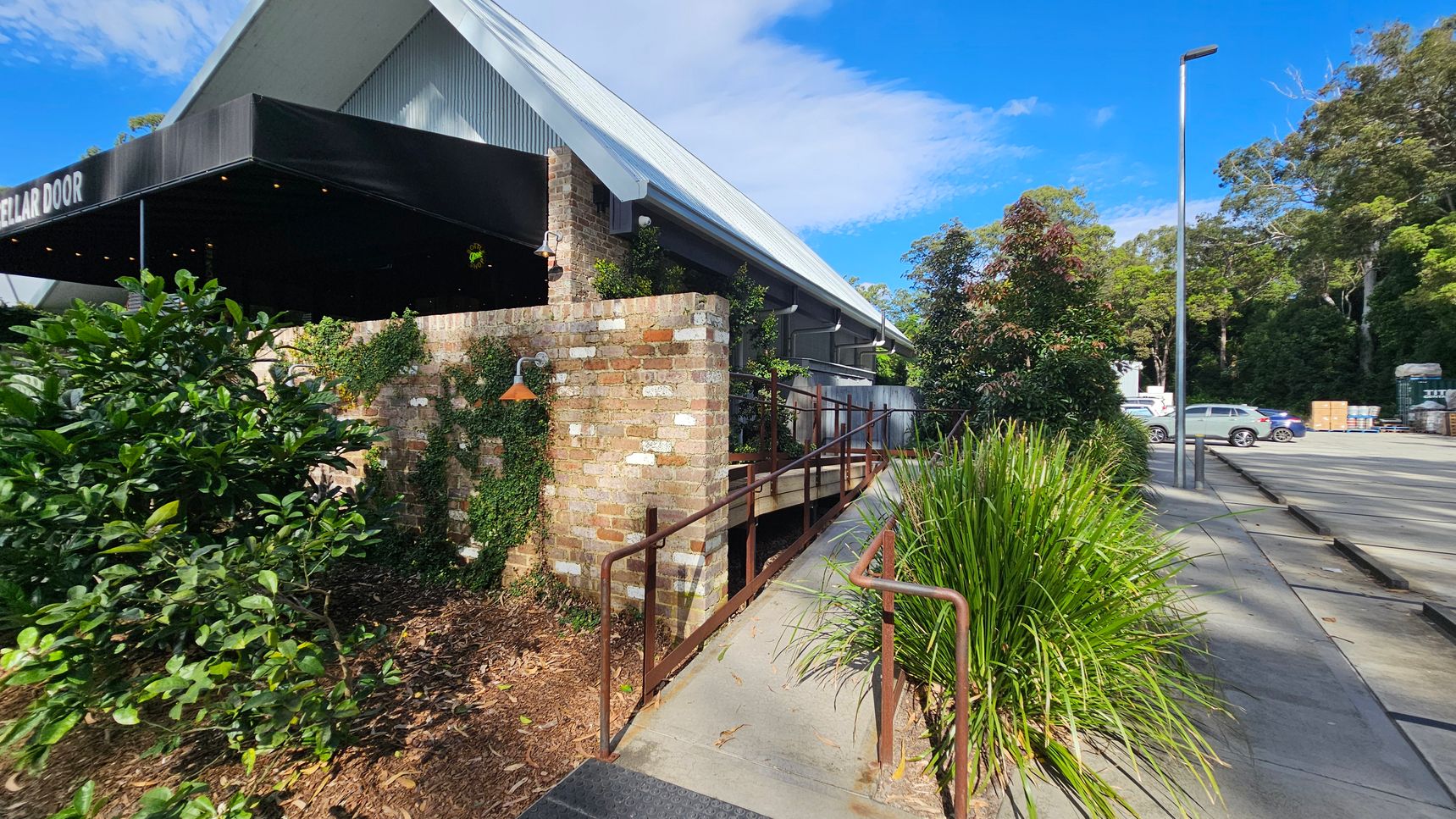 A building with a sloped metal roof and a brick exterior is on the left. A black awning with the words "CELLAR DOOR" extends from the building. A ramp with metal railings leads up to the entrance. Greenery, including bushes and climbing plants, surrounds the area. On the right, a paved area with parked cars and a tall streetlamp is visible. Trees and foliage are in the background. A building with a sloped metal roof and a brick exterior is on the left. A black awning with the words "CELLAR DOOR" extends from the building. A ramp with metal railings leads up to the entrance. Greenery, including bushes and climbing plants, surrounds the area. On the right, a paved area with parked cars and a tall streetlamp is visible. Trees and foliage are in the background.