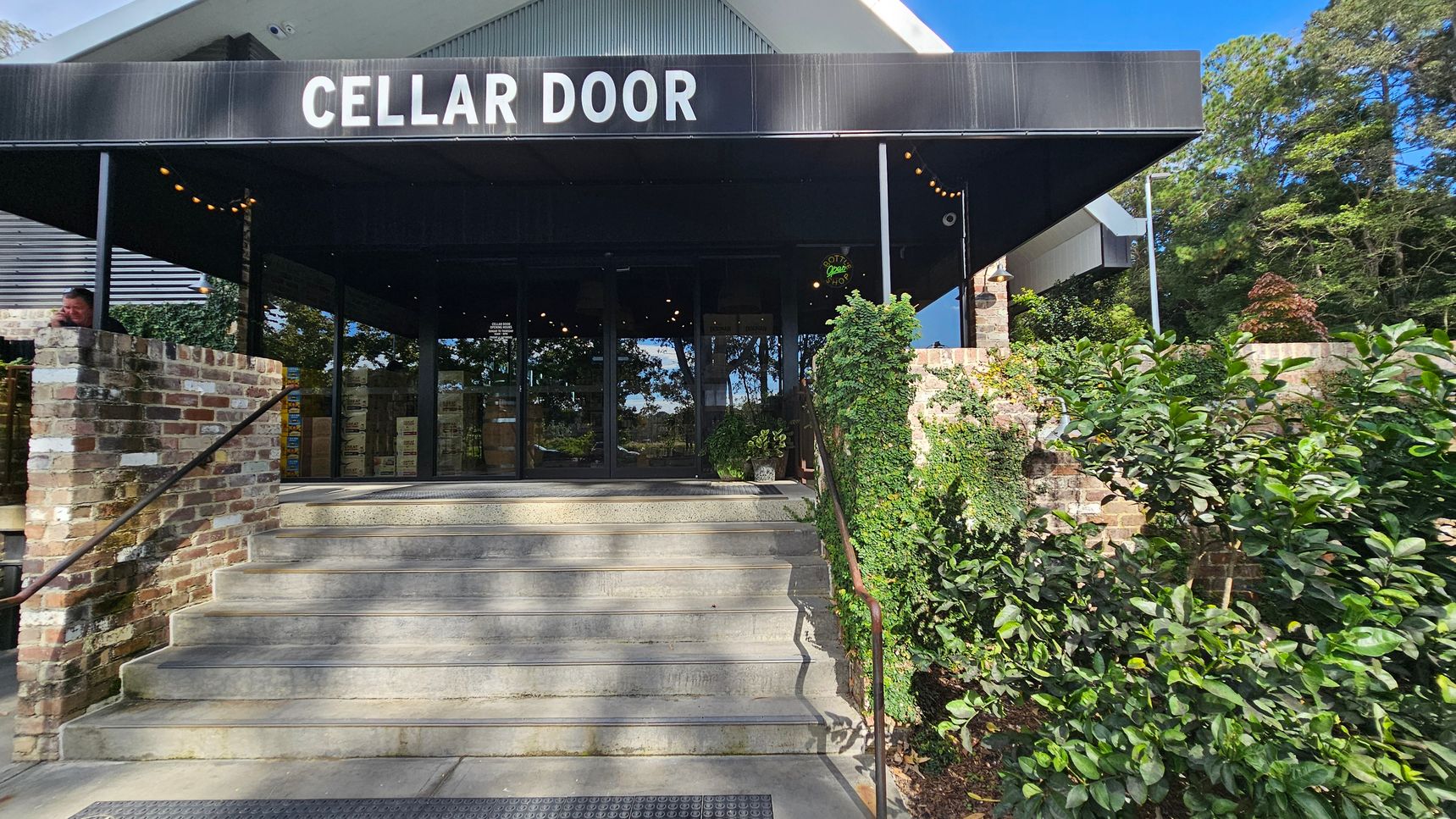 A building entrance with a sign reading "CELLAR DOOR" above. The entrance is accessed by a set of concrete steps flanked by brick walls. On the right, green foliage and vines cover part of the wall. The left side shows a person leaning on the brick wall. The background features glass doors reflecting trees and greenery. The ceiling of the entrance is dark, with string lights visible. A building entrance with a sign reading "CELLAR DOOR" above. The entrance is accessed by a set of concrete steps flanked by brick walls. On the right, green foliage and vines cover part of the wall. The left side shows a person leaning on the brick wall. The background features glass doors reflecting trees and greenery. The ceiling of the entrance is dark, with string lights visible.