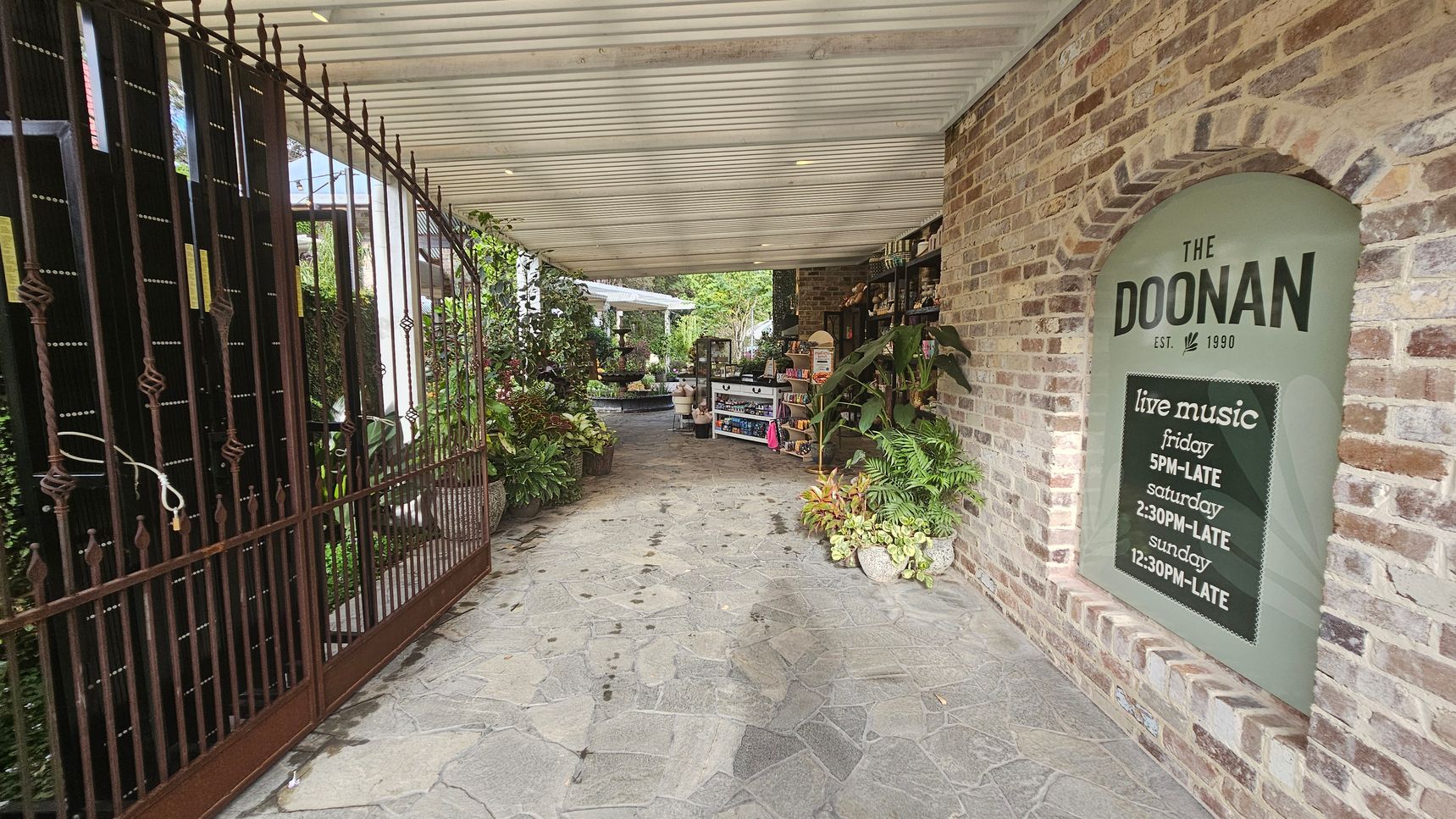 A covered walkway with a stone floor leads to a garden area. On the left, there's a wrought iron gate with decorative elements. On the right, a brick wall features a sign for "The Doonan" with live music timings. Various plants and greenery are placed along the path, and shelves with items are visible. The ceiling is wooden with recessed lighting. In the background, more plants and a seating area can be seen. A covered walkway with a stone floor leads to a garden area. On the left, there's a wrought iron gate with decorative elements. On the right, a brick wall features a sign for "The Doonan" with live music timings. Various plants and greenery are placed along the path, and shelves with items are visible. The ceiling is wooden with recessed lighting. In the background, more plants and a seating area can be seen.
