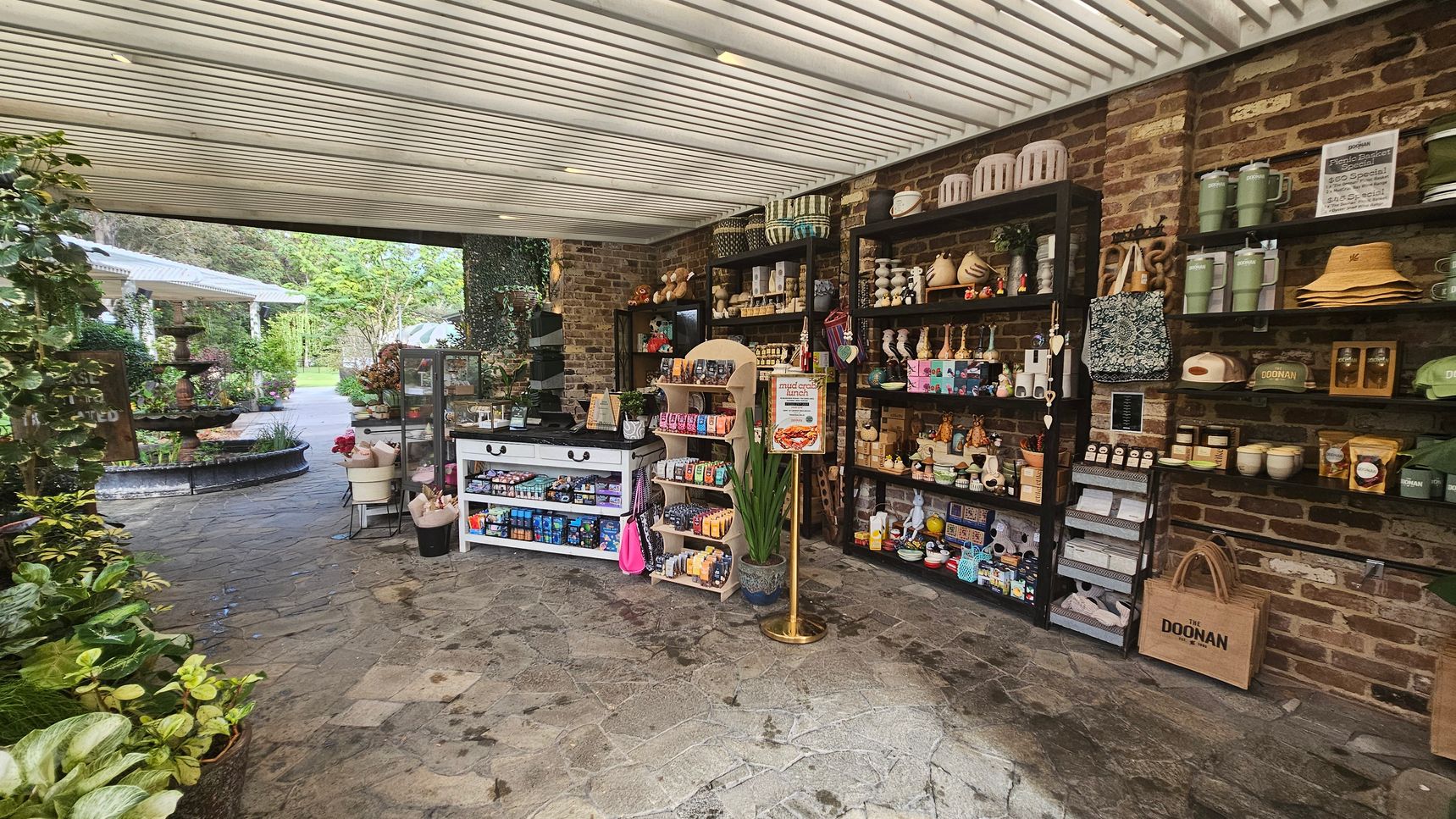 A small shop with a rustic brick wall on the right, featuring shelves filled with various items like mugs, hats, and candles. In the centre, a white display table and a wooden rack hold snacks and small goods. The floor is stone-tiled. On the left, lush green plants and a path lead outside to a garden area with a fountain and more greenery. The ceiling is slatted, allowing natural light to filter through. A small shop with a rustic brick wall on the right, featuring shelves filled with various items like mugs, hats, and candles. In the centre, a white display table and a wooden rack hold snacks and small goods. The floor is stone-tiled. On the left, lush green plants and a path lead outside to a garden area with a fountain and more greenery. The ceiling is slatted, allowing natural light to filter through.