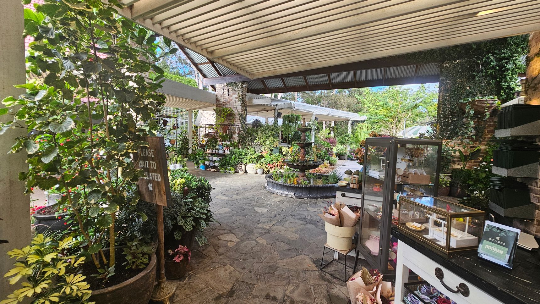 A garden centre with a variety of plants and flowers in pots on the left and centre. A sign reads "Please wait to be seated." In the middle, a stone path leads to a fountain surrounded by more plants. On the right, a display case with items for sale is on a counter, with a sign mentioning gift cards. The ceiling is covered with corrugated metal, and the floor is made of stone tiles. The background features lush greenery and a brick wall. A garden centre with a variety of plants and flowers in pots on the left and centre. A sign reads "Please wait to be seated." In the middle, a stone path leads to a fountain surrounded by more plants. On the right, a display case with items for sale is on a counter, with a sign mentioning gift cards. The ceiling is covered with corrugated metal, and the floor is made of stone tiles. The background features lush greenery and a brick wall.