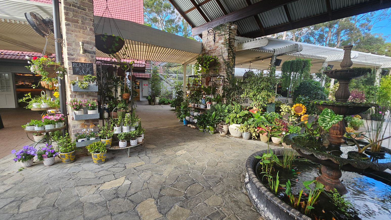 A garden centre with a variety of potted plants displayed on the left, including flowers and greenery. A brick pillar with a "Plants for Sale" sign is adorned with hanging plants. In the centre, more plants are arranged on shelves. On the right, a tiered fountain with water plants is visible. The floor is made of stone tiles, and the ceiling is corrugated metal. In the background, there are more plants and a sign for a pizzeria. A garden centre with a variety of potted plants displayed on the left, including flowers and greenery. A brick pillar with a "Plants for Sale" sign is adorned with hanging plants. In the centre, more plants are arranged on shelves. On the right, a tiered fountain with water plants is visible. The floor is made of stone tiles, and the ceiling is corrugated metal. In the background, there are more plants and a sign for a pizzeria.
