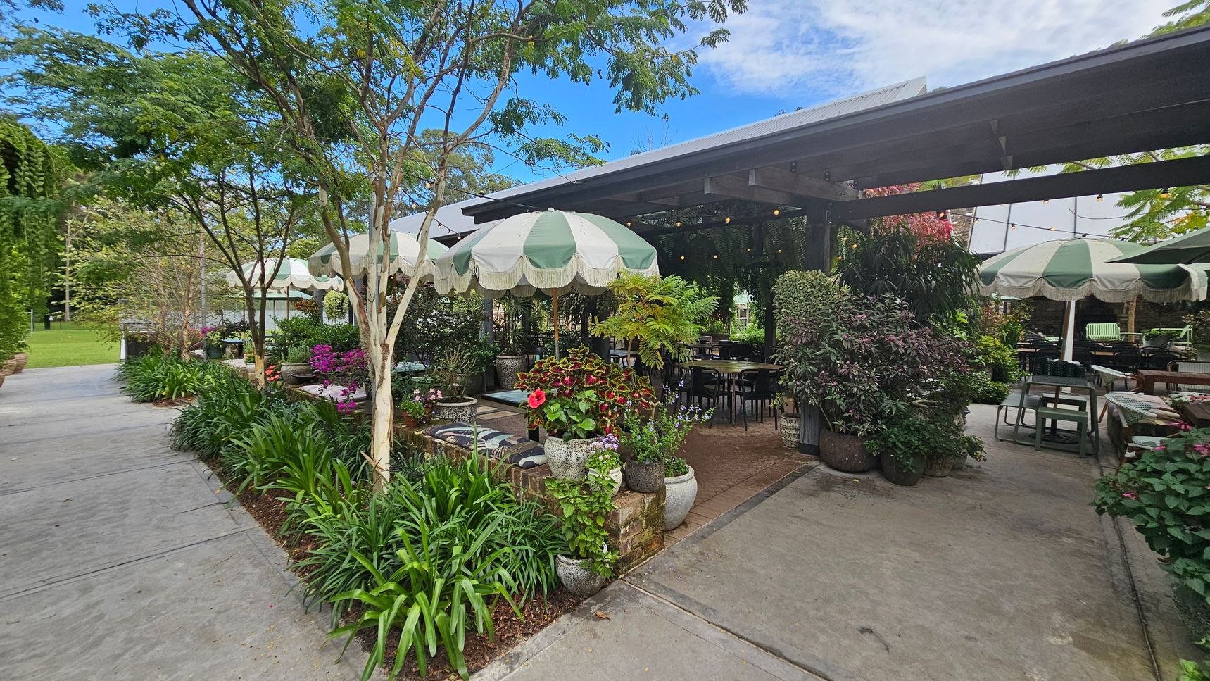 A garden café with green and white striped umbrellas shading tables and chairs. Lush greenery and colourful flowers in pots surround the seating area. A concrete path runs along the left side, bordered by plants and trees. The café structure has a metal roof, and string lights are visible under the canopy. The setting is open and inviting, with a mix of natural and man-made elements. A garden café with green and white striped umbrellas shading tables and chairs. Lush greenery and colourful flowers in pots surround the seating area. A concrete path runs along the left side, bordered by plants and trees. The café structure has a metal roof, and string lights are visible under the canopy. The setting is open and inviting, with a mix of natural and man-made elements.