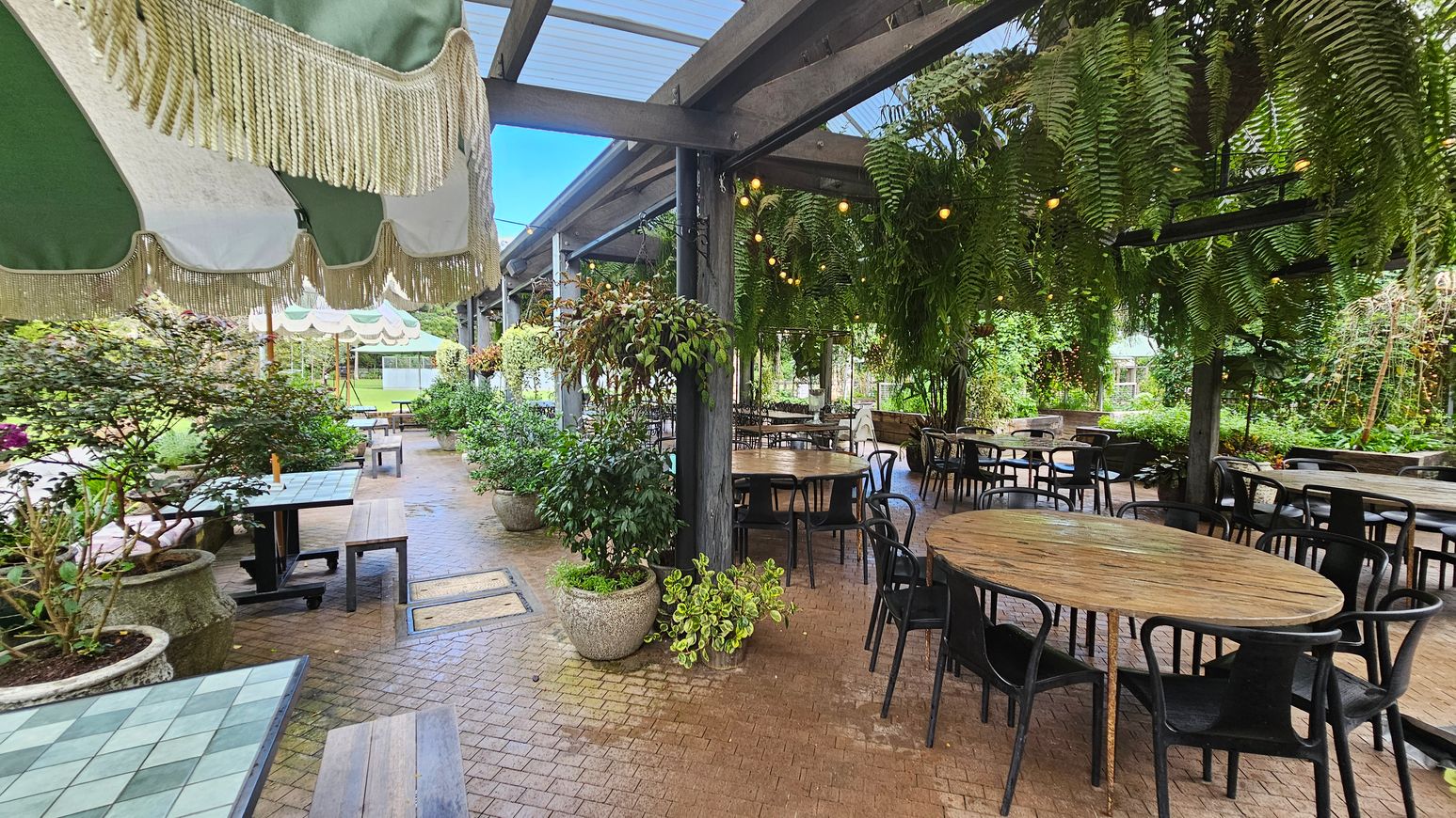 A covered outdoor seating area with round wooden tables and black chairs on the right. Hanging ferns and string lights adorn the ceiling. On the left, there are square tables with green and white checkered tops, accompanied by benches. Large potted plants are scattered throughout the space. The floor is paved with small bricks, and green and white striped awnings with fringe hang above. The background features lush greenery and more seating. A covered outdoor seating area with round wooden tables and black chairs on the right. Hanging ferns and string lights adorn the ceiling. On the left, there are square tables with green and white checkered tops, accompanied by benches. Large potted plants are scattered throughout the space. The floor is paved with small bricks, and green and white striped awnings with fringe hang above. The background features lush greenery and more seating.