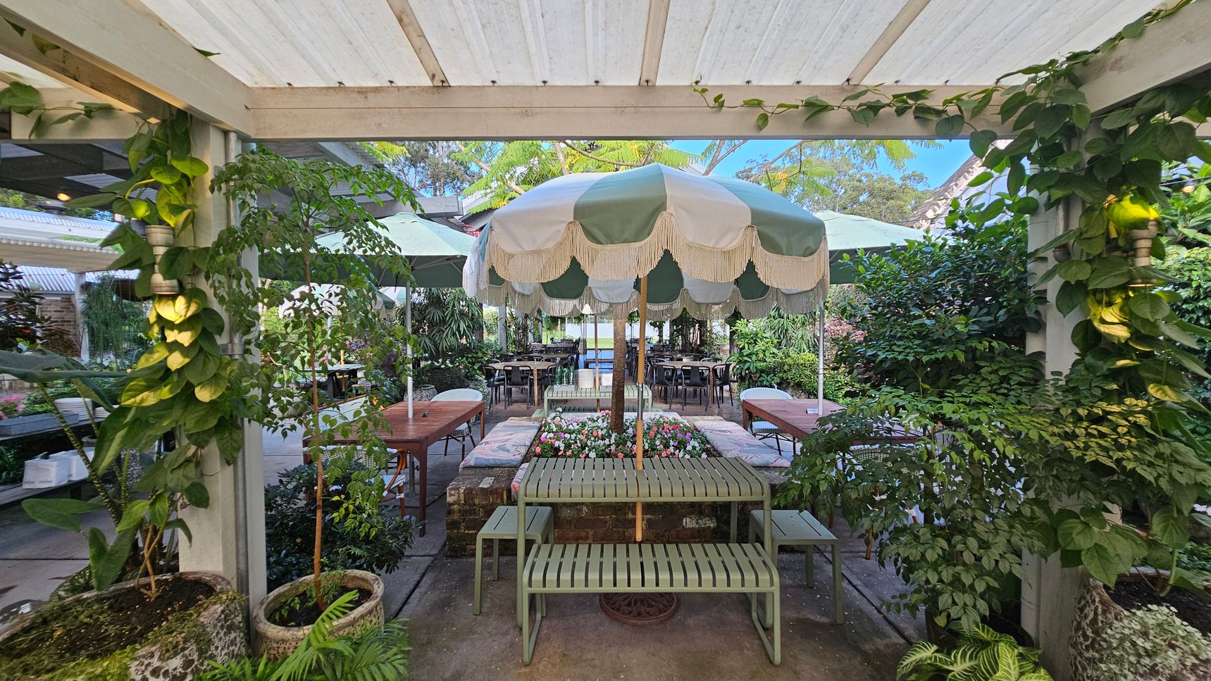 A garden setting with a central green and white striped umbrella shading a bench and table. Surrounding the area are lush green plants and foliage. On either side, there are wooden tables with chairs. The ceiling is a white pergola with climbing plants. The floor is paved, and there are planters with various plants around the space, creating a cosy, natural atmosphere. A garden setting with a central green and white striped umbrella shading a bench and table. Surrounding the area are lush green plants and foliage. On either side, there are wooden tables with chairs. The ceiling is a white pergola with climbing plants. The floor is paved, and there are planters with various plants around the space, creating a cosy, natural atmosphere.
