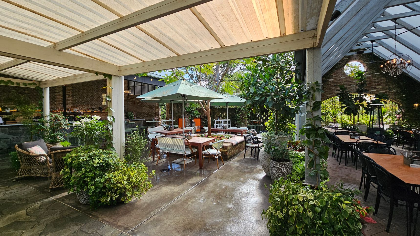 A spacious outdoor patio with a mix of seating arrangements. On the left, there are wicker chairs with cushions and lush green plants in pots. In the centre, a wooden table is surrounded by chairs and benches, shaded by large green umbrellas. The right side features a long table with black chairs under a slanted glass roof. The background has a brick wall with a circular window and more greenery. The floor is a mix of stone and brick, and the ceiling is partially covered with translucent panels. A spacious outdoor patio with a mix of seating arrangements. On the left, there are wicker chairs with cushions and lush green plants in pots. In the centre, a wooden table is surrounded by chairs and benches, shaded by large green umbrellas. The right side features a long table with black chairs under a slanted glass roof. The background has a brick wall with a circular window and more greenery. The floor is a mix of stone and brick, and the ceiling is partially covered with translucent panels.