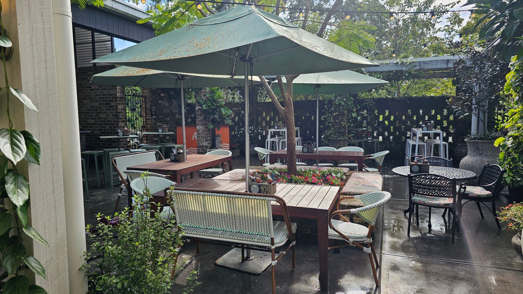 Outdoor seating area with several wooden tables and chairs, some with woven designs. Large green umbrellas provide shade. A variety of plants and flowers are arranged around the space, including a tree in the centre. The ground is paved, and string lights are strung overhead. A brick wall and a wooden structure are in the background, with additional seating and a few stools visible. A large planter is on the right. Outdoor seating area with several wooden tables and chairs, some with woven designs. Large green umbrellas provide shade. A variety of plants and flowers are arranged around the space, including a tree in the centre. The ground is paved, and string lights are strung overhead. A brick wall and a wooden structure are in the background, with additional seating and a few stools visible. A large planter is on the right.