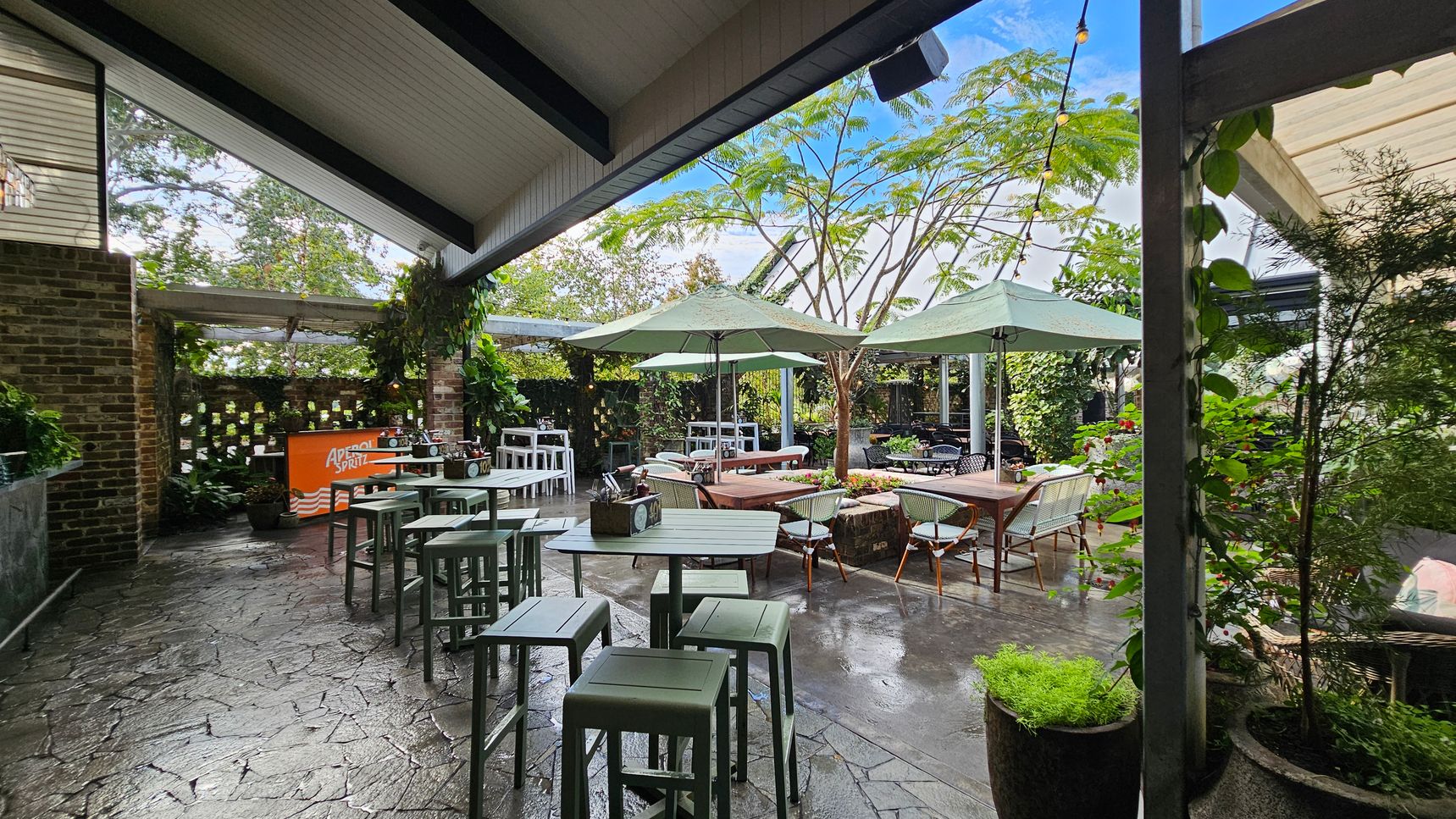 Outdoor seating area with a stone floor and a slanted ceiling. On the left, there's a brick wall with a small bar featuring an "Aperol Spritz" sign. Several high tables with stools are arranged nearby. In the centre, wooden tables with chairs are shaded by large green umbrellas. Lush greenery and potted plants surround the space, with trees visible in the background. String lights are hung across the area, adding a decorative touch. Outdoor seating area with a stone floor and a slanted ceiling. On the left, there's a brick wall with a small bar featuring an "Aperol Spritz" sign. Several high tables with stools are arranged nearby. In the centre, wooden tables with chairs are shaded by large green umbrellas. Lush greenery and potted plants surround the space, with trees visible in the background. String lights are hung across the area, adding a decorative touch.