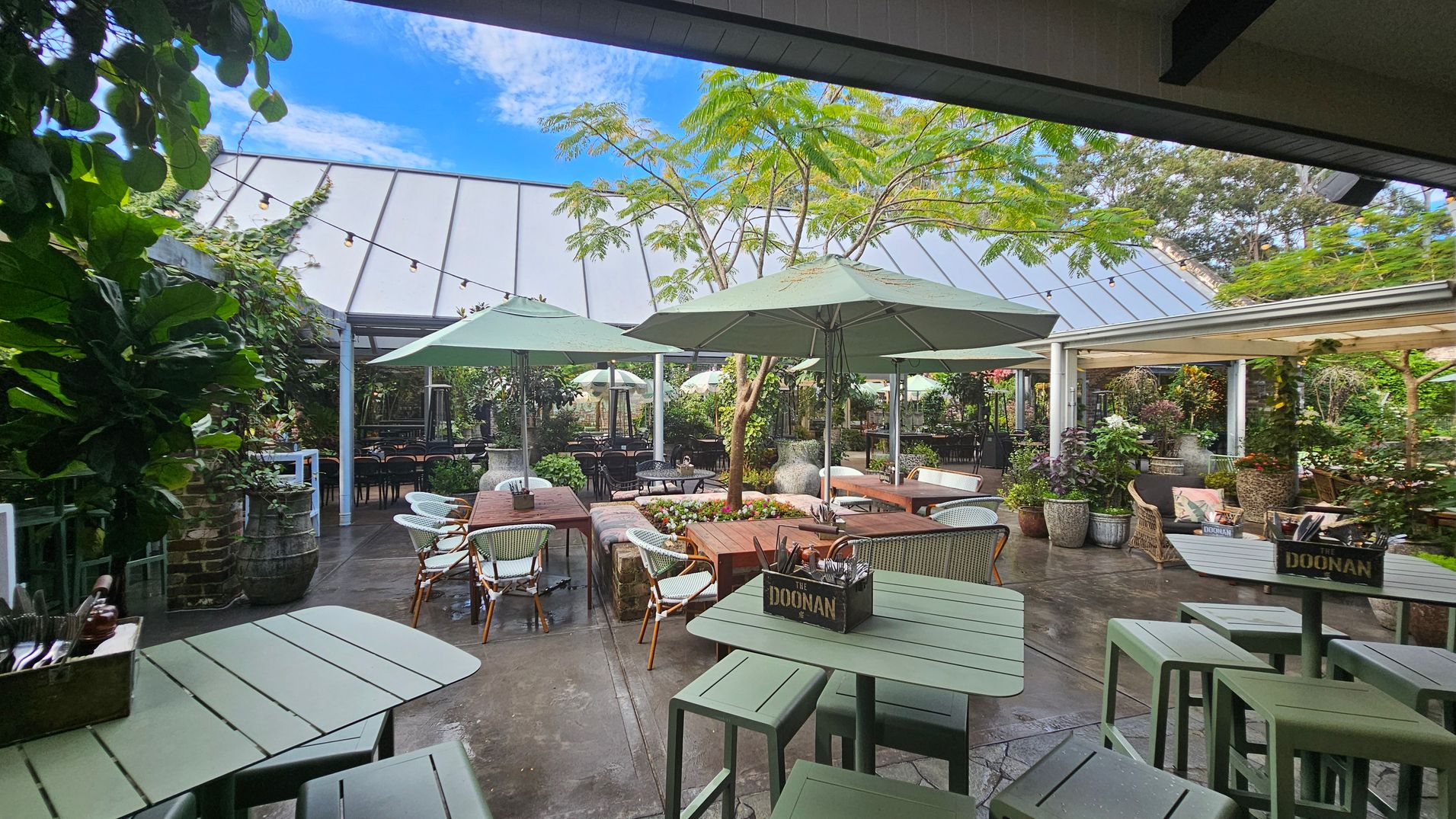 Outdoor seating area with green tables and chairs, surrounded by lush plants. Large umbrellas provide shade over wooden tables. A tree is centrally located, with string lights overhead. The background features a glass-roofed structure and more greenery. The floor is paved, and there are decorative planters throughout. Outdoor seating area with green tables and chairs, surrounded by lush plants. Large umbrellas provide shade over wooden tables. A tree is centrally located, with string lights overhead. The background features a glass-roofed structure and more greenery. The floor is paved, and there are decorative planters throughout.
