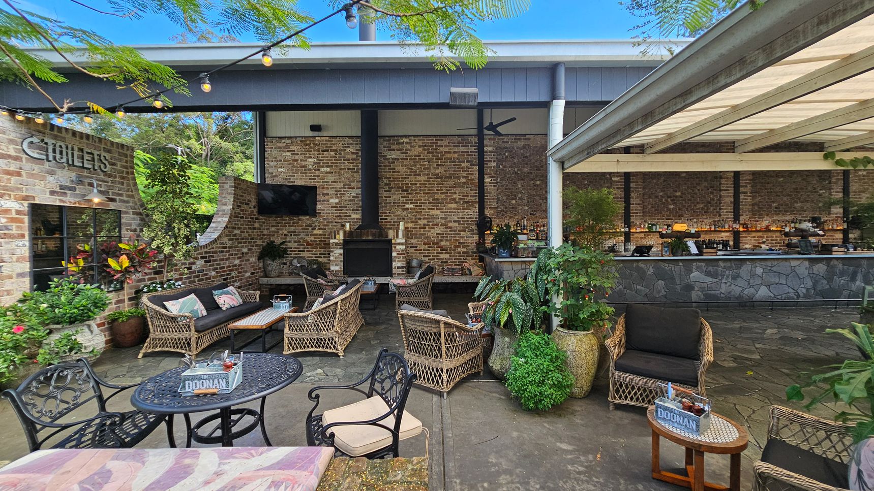 Outdoor seating area with wicker furniture and a brick wall backdrop. On the left, a sign for toilets is visible on the brick wall, surrounded by potted plants. In the centre, a fireplace is flanked by a TV and more seating. On the right, a bar with a stone facade is under a covered area. String lights hang above, and the floor is paved with stone tiles. Outdoor seating area with wicker furniture and a brick wall backdrop. On the left, a sign for toilets is visible on the brick wall, surrounded by potted plants. In the centre, a fireplace is flanked by a TV and more seating. On the right, a bar with a stone facade is under a covered area. String lights hang above, and the floor is paved with stone tiles.