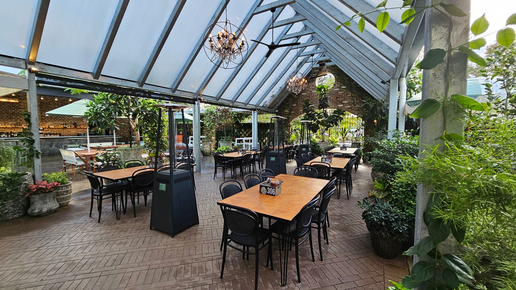 A spacious outdoor dining area with a glass roof and brick flooring. In the centre, there are several wooden tables with black chairs, each marked with a number. On the left, a bar area with a brick wall and a large umbrella. On the right, lush greenery and potted plants line the space. Hanging chandeliers and ceiling fans are suspended from the roof. The atmosphere is open and inviting, with a mix of natural and industrial elements. A spacious outdoor dining area with a glass roof and brick flooring. In the centre, there are several wooden tables with black chairs, each marked with a number. On the left, a bar area with a brick wall and a large umbrella. On the right, lush greenery and potted plants line the space. Hanging chandeliers and ceiling fans are suspended from the roof. The atmosphere is open and inviting, with a mix of natural and industrial elements.