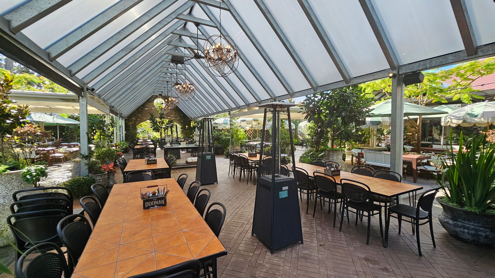 A spacious outdoor dining area with a glass roof supported by metal beams. Long wooden tables with black chairs are arranged in rows. Two tall outdoor heaters are positioned along the centre aisle. The floor is paved with bricks in a herringbone pattern. Lush greenery and potted plants surround the space, with additional seating visible in the background under green umbrellas. Chandeliers hang from the ceiling, adding a decorative touch. A spacious outdoor dining area with a glass roof supported by metal beams. Long wooden tables with black chairs are arranged in rows. Two tall outdoor heaters are positioned along the centre aisle. The floor is paved with bricks in a herringbone pattern. Lush greenery and potted plants surround the space, with additional seating visible in the background under green umbrellas. Chandeliers hang from the ceiling, adding a decorative touch.