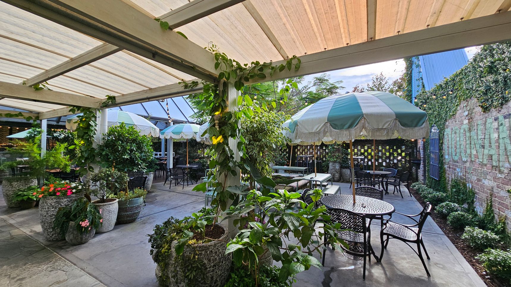 Outdoor patio with a covered pergola, featuring climbing plants on the beams. Several large potted plants with vibrant flowers are scattered around. Black metal tables and chairs are arranged under green and white striped umbrellas. A brick wall in the background is partially covered with ivy, and string lights are hung above. The floor is paved with stone tiles. Outdoor patio with a covered pergola, featuring climbing plants on the beams. Several large potted plants with vibrant flowers are scattered around. Black metal tables and chairs are arranged under green and white striped umbrellas. A brick wall in the background is partially covered with ivy, and string lights are hung above. The floor is paved with stone tiles.