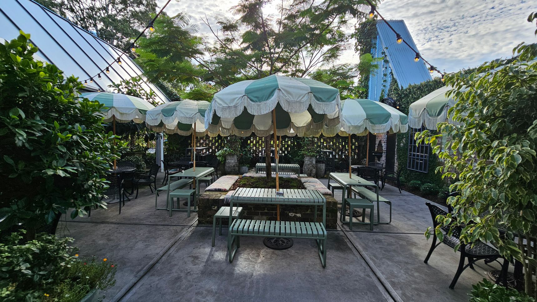 Outdoor seating area with green and white striped umbrellas in the centre, surrounded by green metal benches and tables. The ground is concrete, and there are string lights overhead. Lush greenery and plants border the area on the left and right, with a blue-roofed structure visible in the background. The setting has a garden-like atmosphere with a mix of natural and architectural elements. Outdoor seating area with green and white striped umbrellas in the centre, surrounded by green metal benches and tables. The ground is concrete, and there are string lights overhead. Lush greenery and plants border the area on the left and right, with a blue-roofed structure visible in the background. The setting has a garden-like atmosphere with a mix of natural and architectural elements.
