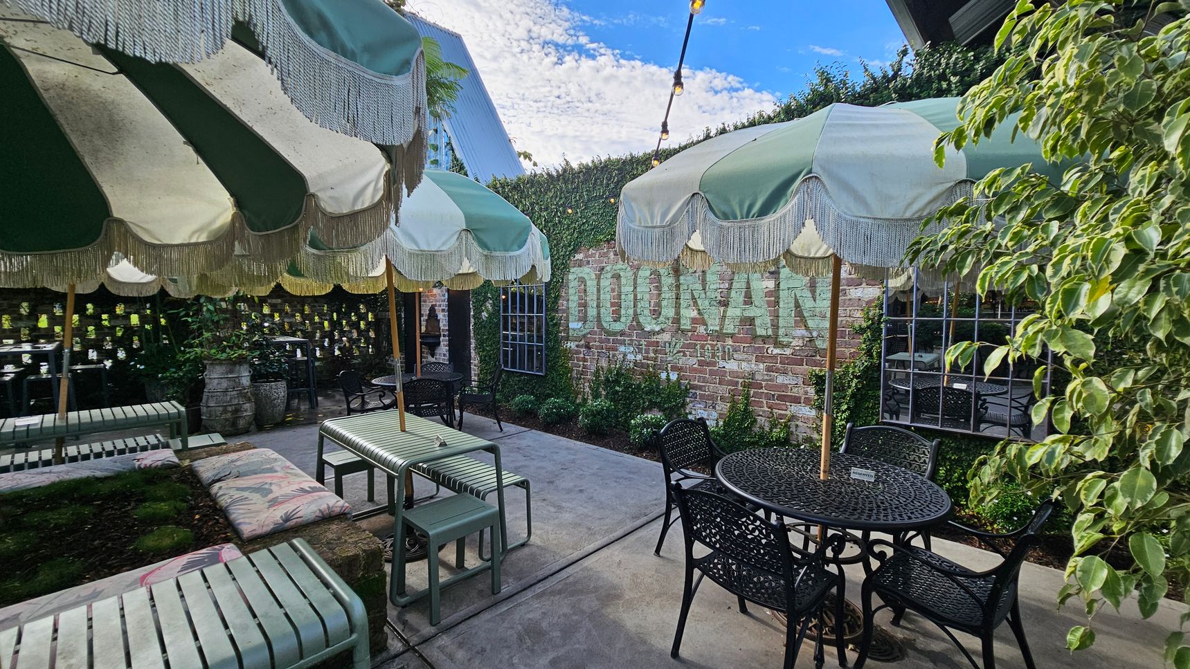 A courtyard with green and white striped umbrellas featuring fringe, providing shade over metal tables and chairs. On the left, a bench with a floral cushion is visible. The background has a brick wall with the word "DOONAN" painted on it, partially covered by ivy. A window with a grid pattern is set into the wall. The right side has a leafy plant, and string lights are strung across the area. The floor is concrete. A courtyard with green and white striped umbrellas featuring fringe, providing shade over metal tables and chairs. On the left, a bench with a floral cushion is visible. The background has a brick wall with the word "DOONAN" painted on it, partially covered by ivy. A window with a grid pattern is set into the wall. The right side has a leafy plant, and string lights are strung across the area. The floor is concrete.