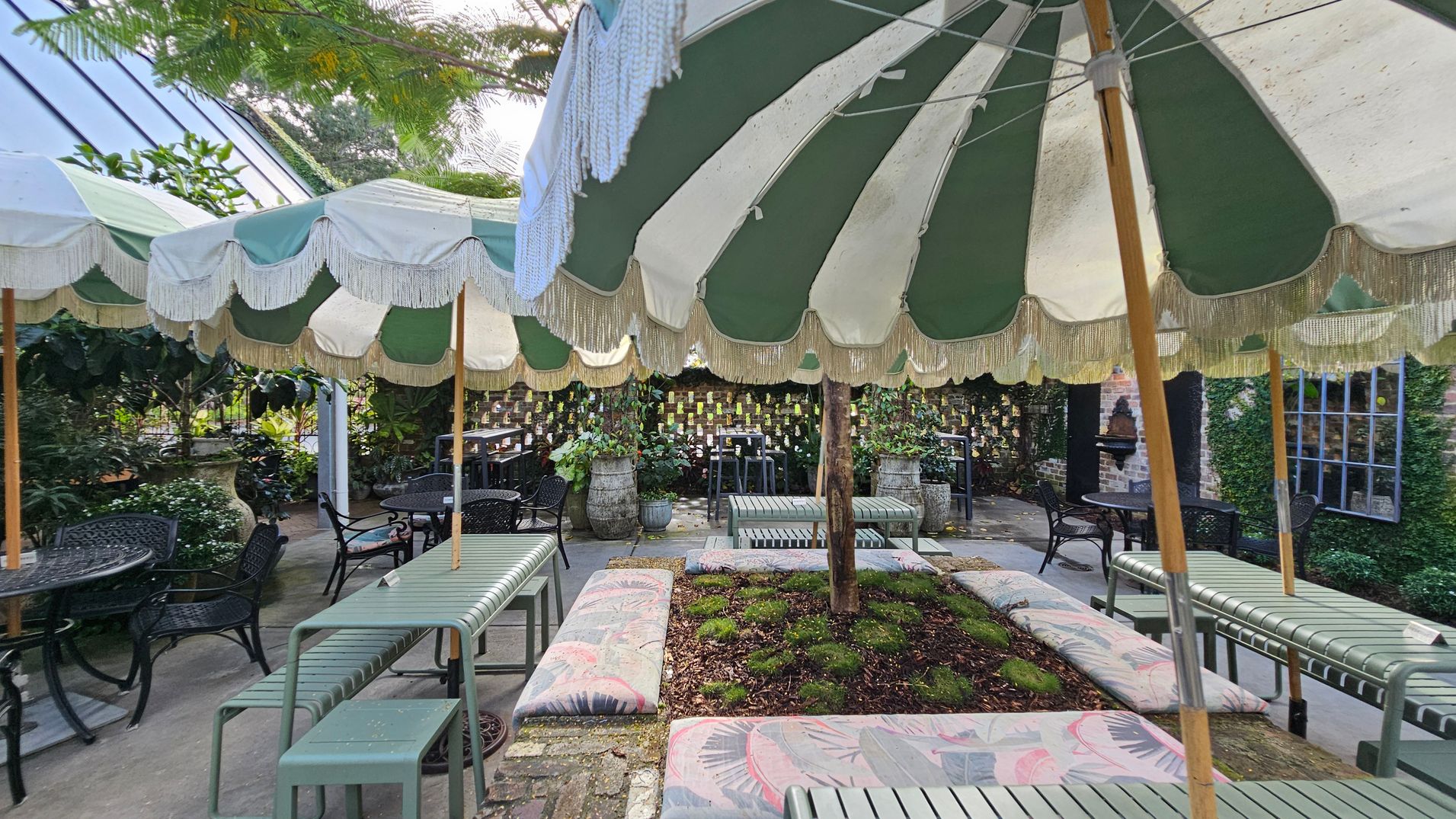 A garden setting with green and white striped parasols featuring fringe, providing shade over seating areas. The seating includes metal benches and cushioned seats around a small garden bed with plants. In the background, there are potted plants and a brick wall adorned with small hanging items. Tables and chairs are scattered throughout, with greenery and foliage visible around the perimeter. The floor is paved with stone tiles. A garden setting with green and white striped parasols featuring fringe, providing shade over seating areas. The seating includes metal benches and cushioned seats around a small garden bed with plants. In the background, there are potted plants and a brick wall adorned with small hanging items. Tables and chairs are scattered throughout, with greenery and foliage visible around the perimeter. The floor is paved with stone tiles.