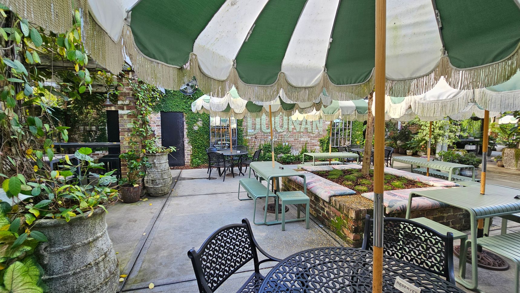 Outdoor seating area with green and white striped umbrellas providing shade. On the left, there are large planters with climbing plants against a brick wall. In the centre, metal tables and benches are arranged around a raised garden bed. The background features a brick wall with ivy and a sign partially visible. The floor is concrete, and the area is surrounded by lush greenery. Outdoor seating area with green and white striped umbrellas providing shade. On the left, there are large planters with climbing plants against a brick wall. In the centre, metal tables and benches are arranged around a raised garden bed. The background features a brick wall with ivy and a sign partially visible. The floor is concrete, and the area is surrounded by lush greenery.