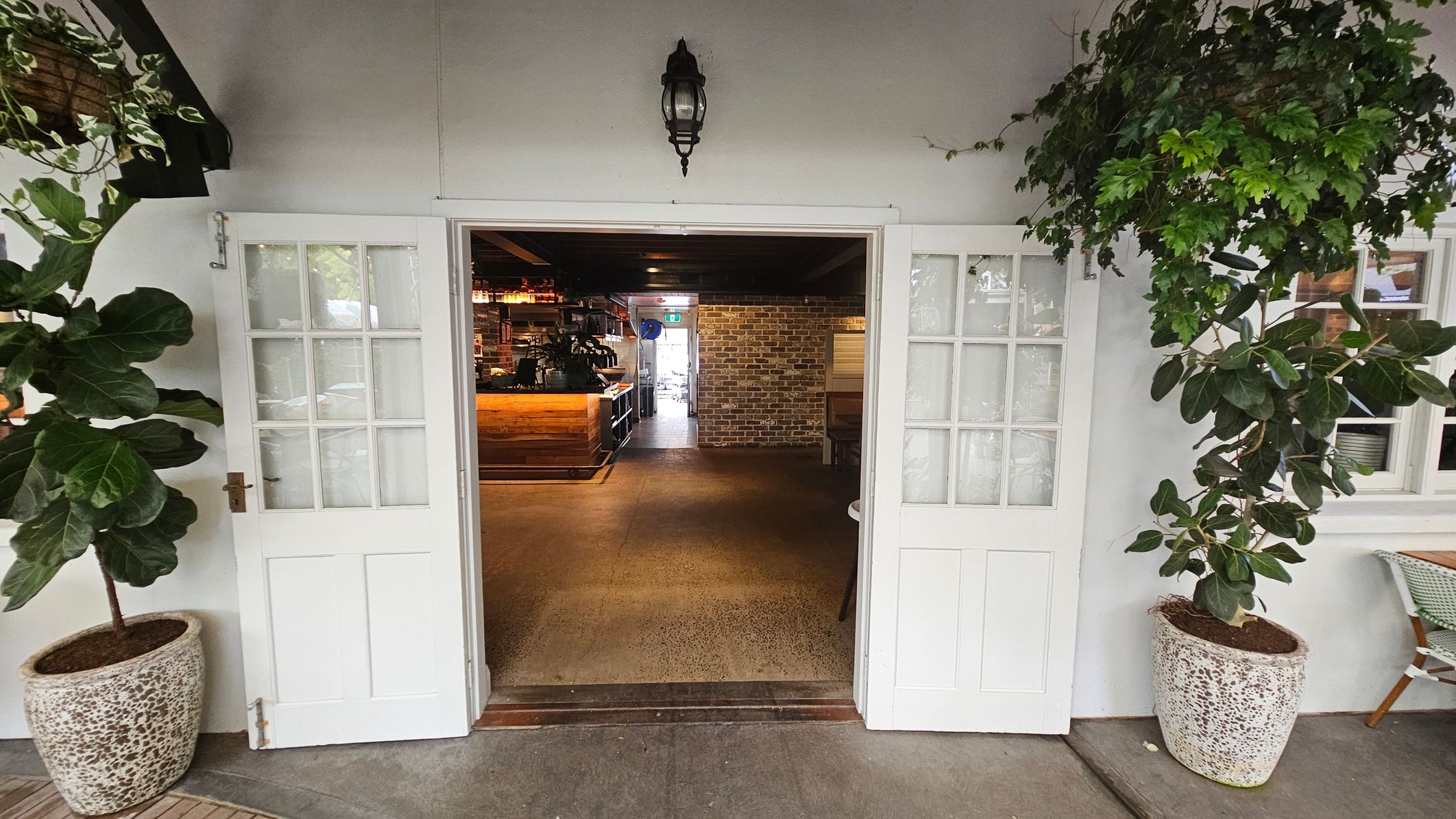 White double doors with glass panes open into an interior space. On either side of the entrance, there are large potted plants with broad green leaves. Inside, a wooden counter is visible to the left, and a brick wall is at the back. The floor is a polished concrete, and a black lantern-style light fixture is mounted above the doors. A small table and chair are partially visible on the right. White double doors with glass panes open into an interior space. On either side of the entrance, there are large potted plants with broad green leaves. Inside, a wooden counter is visible to the left, and a brick wall is at the back. The floor is a polished concrete, and a black lantern-style light fixture is mounted above the doors. A small table and chair are partially visible on the right.