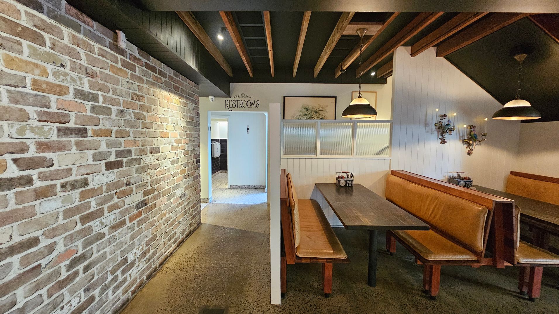 A restaurant interior with a rustic brick wall on the left. In the centre, an entrance marked "Restrooms" leads to a hallway. On the right, there are wooden booths with brown leather seating and dark tables. Above the booths, pendant lights hang from a ceiling with exposed wooden beams. The wall behind the booths is white with decorative sconces. The floor is polished concrete. A restaurant interior with a rustic brick wall on the left. In the centre, an entrance marked "Restrooms" leads to a hallway. On the right, there are wooden booths with brown leather seating and dark tables. Above the booths, pendant lights hang from a ceiling with exposed wooden beams. The wall behind the booths is white with decorative sconces. The floor is polished concrete.