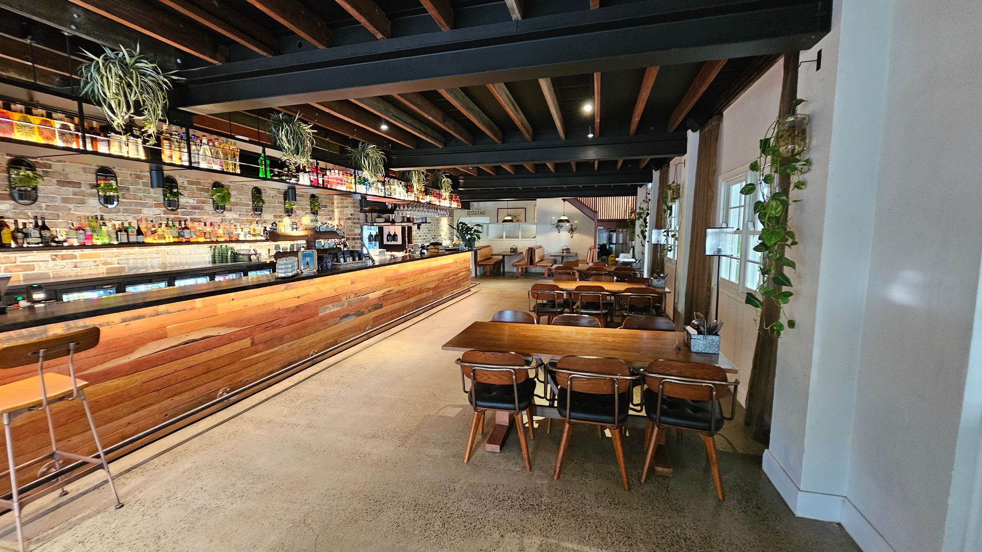 A spacious bar interior with a long wooden counter on the left, lined with various bottles and plants above. The back wall is exposed brick with additional plants and mirrors. On the right, there are wooden tables and chairs, with more plants hanging on the walls. The ceiling features exposed wooden beams, and the floor is polished concrete. In the background, there are more seating areas with booths and pendant lights. A spacious bar interior with a long wooden counter on the left, lined with various bottles and plants above. The back wall is exposed brick with additional plants and mirrors. On the right, there are wooden tables and chairs, with more plants hanging on the walls. The ceiling features exposed wooden beams, and the floor is polished concrete. In the background, there are more seating areas with booths and pendant lights.