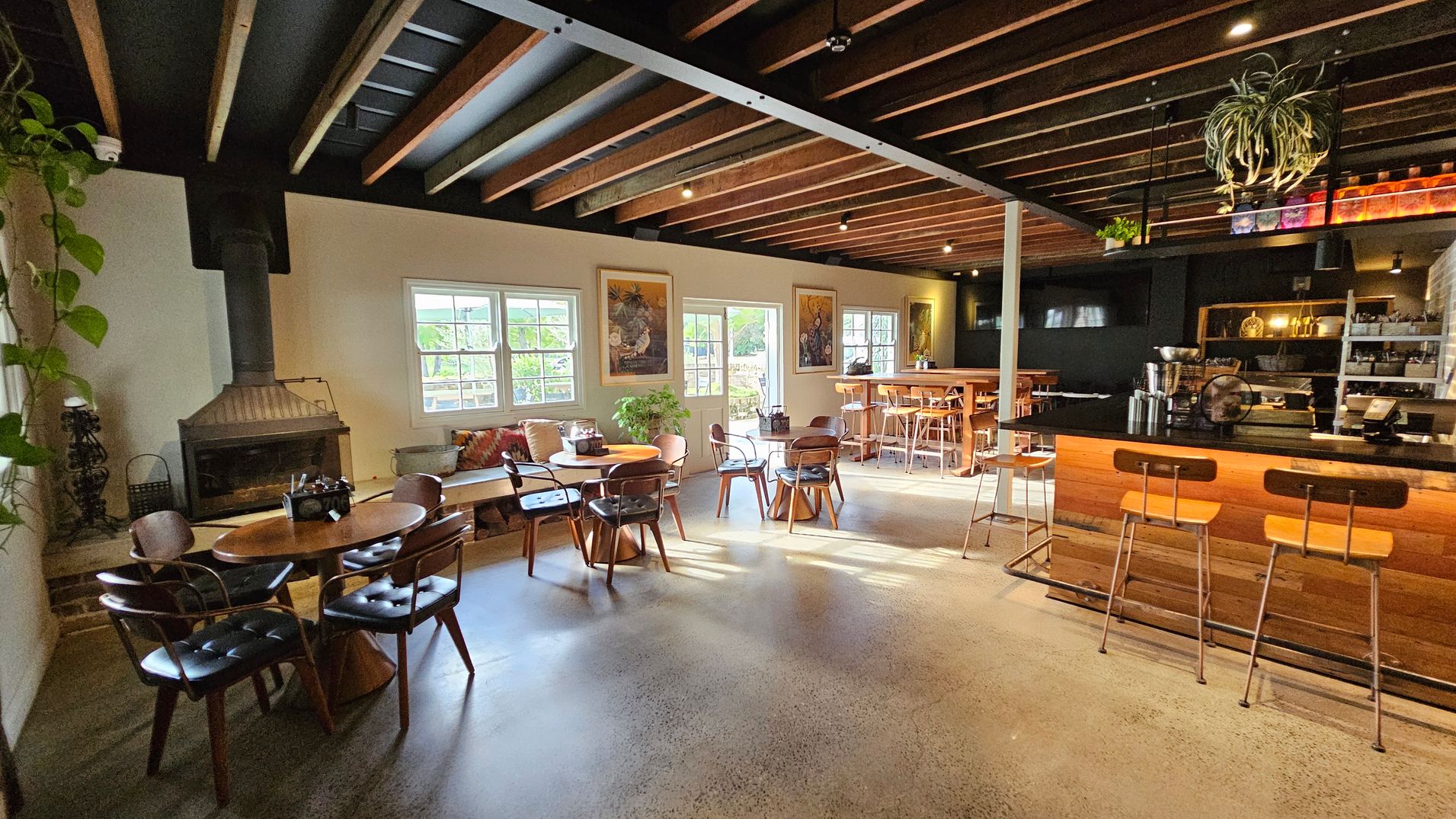 A cosy café interior with a polished concrete floor and a ceiling featuring exposed wooden beams. On the left, there's a fireplace next to a round wooden table with chairs. Large windows allow natural light to illuminate the space. In the centre, more tables and chairs are arranged, with a plant adding greenery. On the right, a wooden bar with stools is visible, with shelves and a coffee machine behind it. A hanging plant is suspended from the ceiling. A cosy café interior with a polished concrete floor and a ceiling featuring exposed wooden beams. On the left, there's a fireplace next to a round wooden table with chairs. Large windows allow natural light to illuminate the space. In the centre, more tables and chairs are arranged, with a plant adding greenery. On the right, a wooden bar with stools is visible, with shelves and a coffee machine behind it. A hanging plant is suspended from the ceiling.