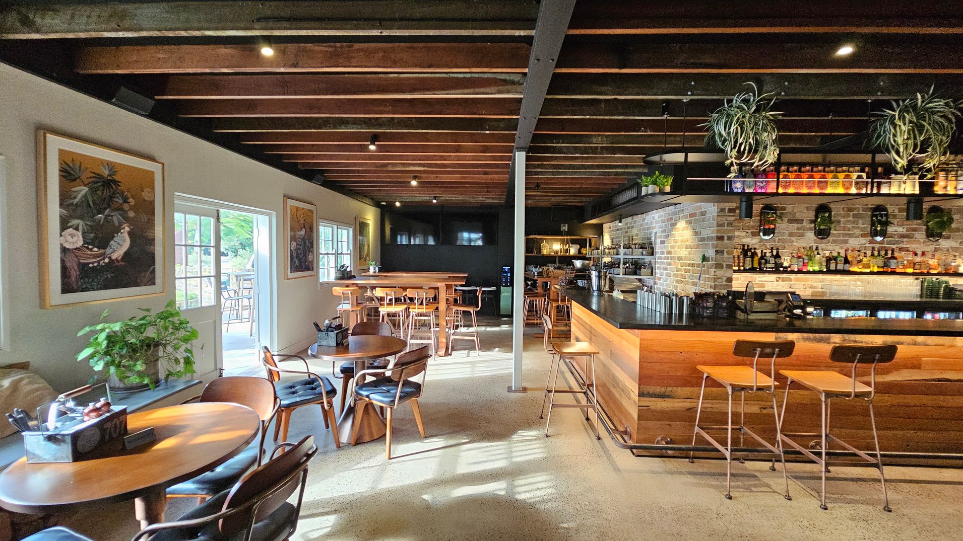 A cosy café interior with wooden beams on the ceiling. On the left, there are round wooden tables with chairs, a plant, and framed artwork on the wall. Sunlight streams through a door leading outside. In the middle, more tables and chairs are arranged. On the right, a bar with a brick wall backdrop displays various bottles, with plants hanging above. The floor is polished concrete, and the atmosphere is warm and inviting. A cosy café interior with wooden beams on the ceiling. On the left, there are round wooden tables with chairs, a plant, and framed artwork on the wall. Sunlight streams through a door leading outside. In the middle, more tables and chairs are arranged. On the right, a bar with a brick wall backdrop displays various bottles, with plants hanging above. The floor is polished concrete, and the atmosphere is warm and inviting.