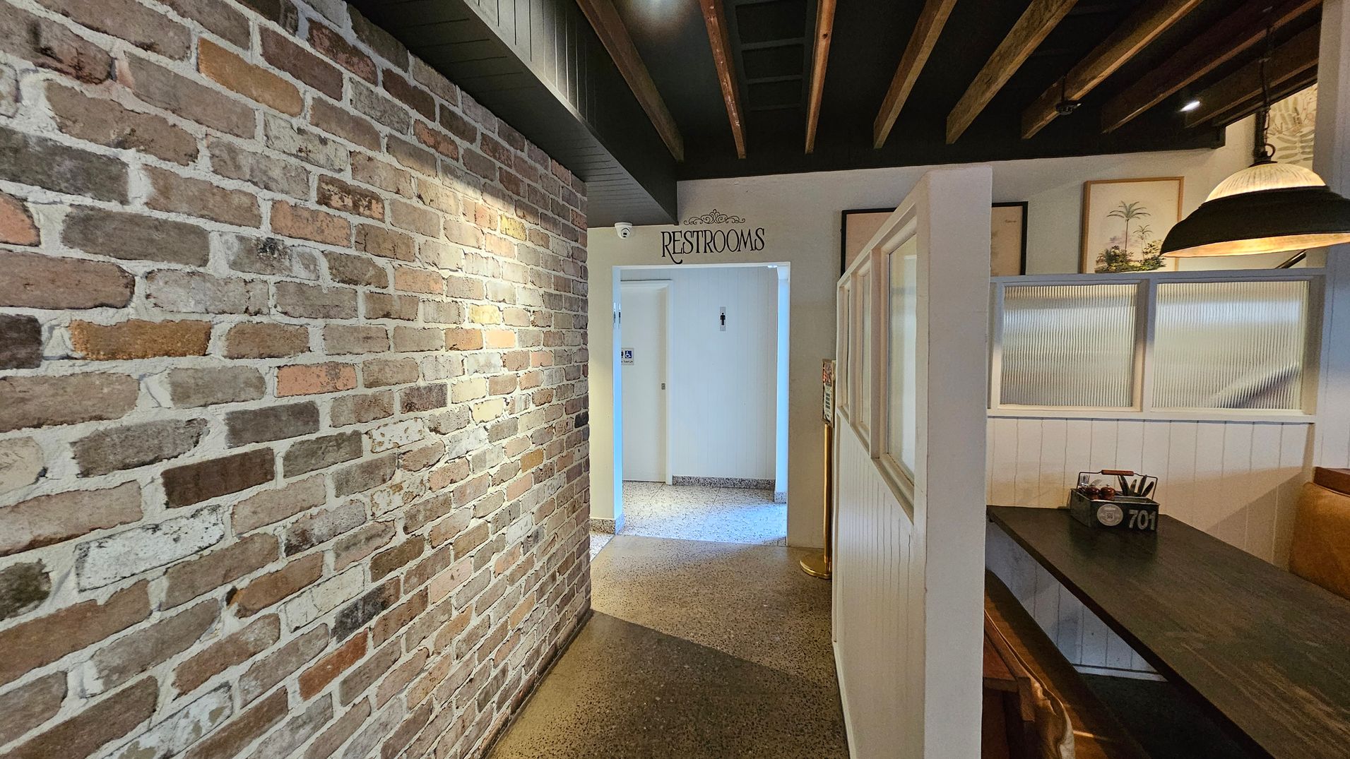 A hallway with a rustic brick wall on the left and a polished stone floor. On the right, there is a dining booth with a dark wooden table and a partition with frosted glass panels. Above the booth, a large pendant light hangs from the ceiling, which features exposed wooden beams. In the centre, a doorway leads to restrooms, marked by a sign above. The walls near the restrooms are painted white, and there are framed pictures on the wall near the booth. A hallway with a rustic brick wall on the left and a polished stone floor. On the right, there is a dining booth with a dark wooden table and a partition with frosted glass panels. Above the booth, a large pendant light hangs from the ceiling, which features exposed wooden beams. In the centre, a doorway leads to restrooms, marked by a sign above. The walls near the restrooms are painted white, and there are framed pictures on the wall near the booth.