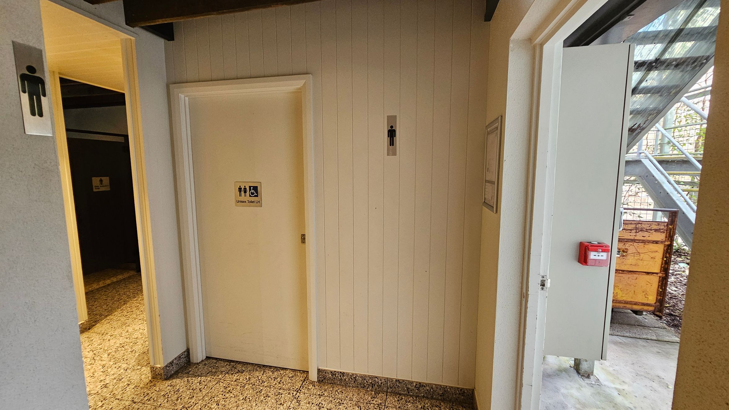 A corridor with a tiled floor and a white panelled wall. On the left, there is a door with a male restroom sign. In the centre, another door has a sign indicating a unisex and accessible toilet. On the right, an open door leads outside, revealing a metal staircase and a rusty container. A red fire alarm is mounted on the wall near the open door. The ceiling has exposed beams. A corridor with a tiled floor and a white panelled wall. On the left, there is a door with a male restroom sign. In the centre, another door has a sign indicating a unisex and accessible toilet. On the right, an open door leads outside, revealing a metal staircase and a rusty container. A red fire alarm is mounted on the wall near the open door. The ceiling has exposed beams.