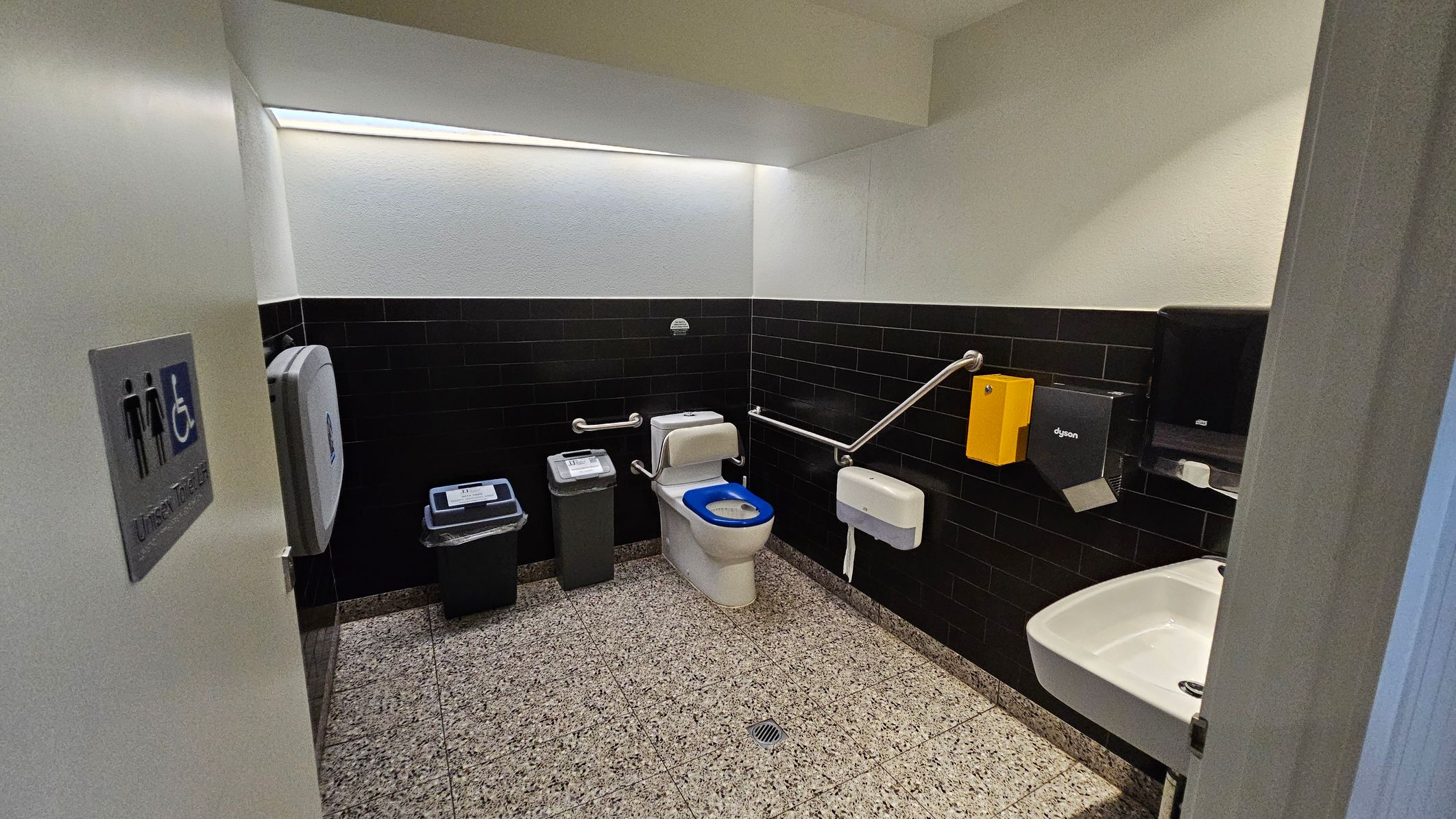 A public accessible toilet with a white toilet featuring a blue seat cover in the centre. To the left, there is a bin and a baby changing station. The walls are tiled with black tiles halfway up, and the floor is speckled grey. On the right, a white sink is mounted on the wall with a hand dryer and paper towel dispenser above it. Stainless steel grab bars are installed around the toilet. The ceiling is plain white, and a sign on the door indicates it is a unisex toilet. A public accessible toilet with a white toilet featuring a blue seat cover in the centre. To the left, there is a bin and a baby changing station. The walls are tiled with black tiles halfway up, and the floor is speckled grey. On the right, a white sink is mounted on the wall with a hand dryer and paper towel dispenser above it. Stainless steel grab bars are installed around the toilet. The ceiling is plain white, and a sign on the door indicates it is a unisex toilet.