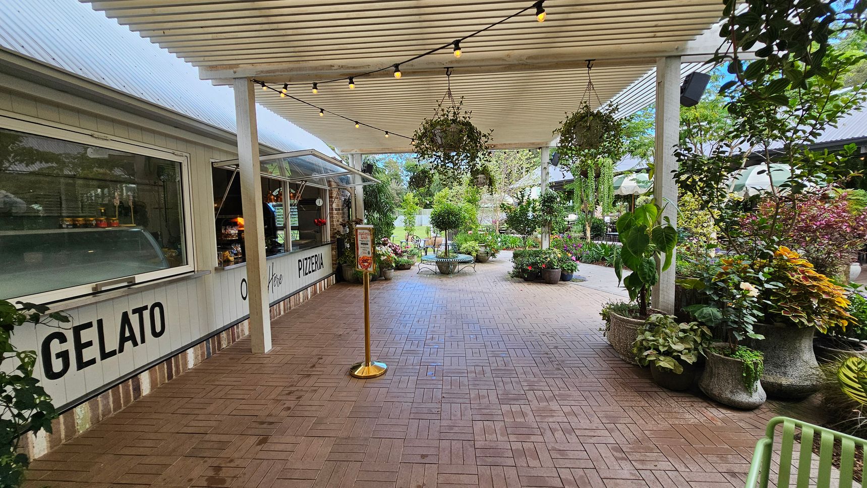 A covered outdoor area with a gelato and pizzeria counter on the left, featuring a white facade and large windows. The ceiling has a slatted design with string lights and hanging plants. The floor is paved with interlocking bricks. On the right, there are various potted plants and greenery. In the background, more plants and a seating area with a bench are visible, creating a garden-like atmosphere. A covered outdoor area with a gelato and pizzeria counter on the left, featuring a white facade and large windows. The ceiling has a slatted design with string lights and hanging plants. The floor is paved with interlocking bricks. On the right, there are various potted plants and greenery. In the background, more plants and a seating area with a bench are visible, creating a garden-like atmosphere.