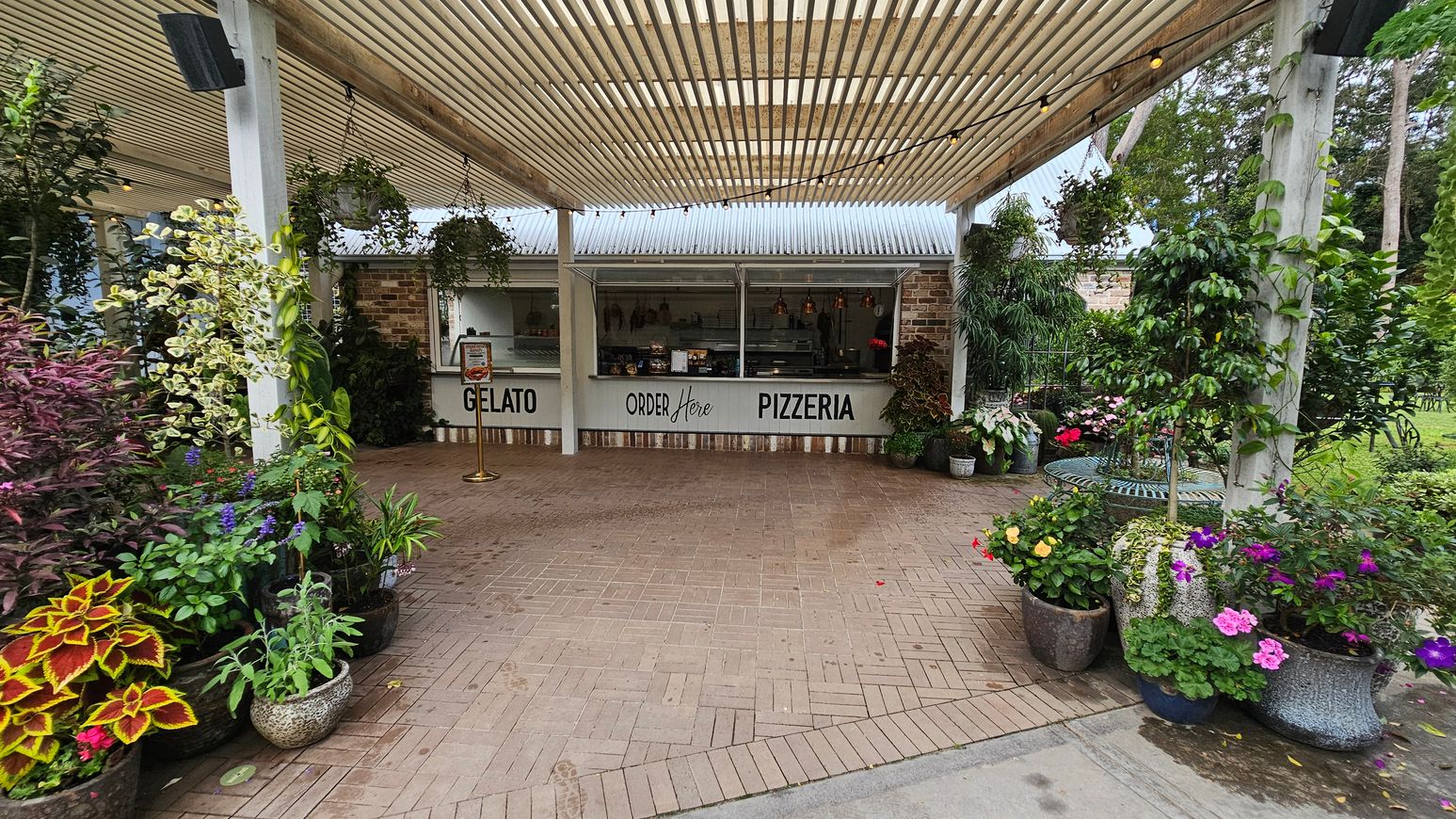 A pizzeria and gelato stand with a sign reading "Order Here" is in the centre, featuring a brick wall and a corrugated metal roof. The foreground has a paved area with a variety of potted plants and flowers on both sides, including colourful blooms and lush greenery. The ceiling is a slatted pergola with string lights. In the background, there are more plants and trees, creating a garden-like atmosphere. A pizzeria and gelato stand with a sign reading "Order Here" is in the centre, featuring a brick wall and a corrugated metal roof. The foreground has a paved area with a variety of potted plants and flowers on both sides, including colourful blooms and lush greenery. The ceiling is a slatted pergola with string lights. In the background, there are more plants and trees, creating a garden-like atmosphere.