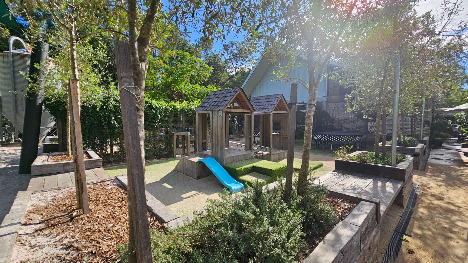 A children's play area with a wooden structure featuring two small peaked roofs and a blue slide in the centre. The ground is covered with sand and bordered by low stone walls. Trees and shrubs are planted around the area, providing shade and greenery. In the background, there is a large building with a corrugated metal exterior. To the left, a tall metal structure is partially visible. The area is well-maintained and designed for outdoor play. A children's play area with a wooden structure featuring two small peaked roofs and a blue slide in the centre. The ground is covered with sand and bordered by low stone walls. Trees and shrubs are planted around the area, providing shade and greenery. In the background, there is a large building with a corrugated metal exterior. To the left, a tall metal structure is partially visible. The area is well-maintained and designed for outdoor play.