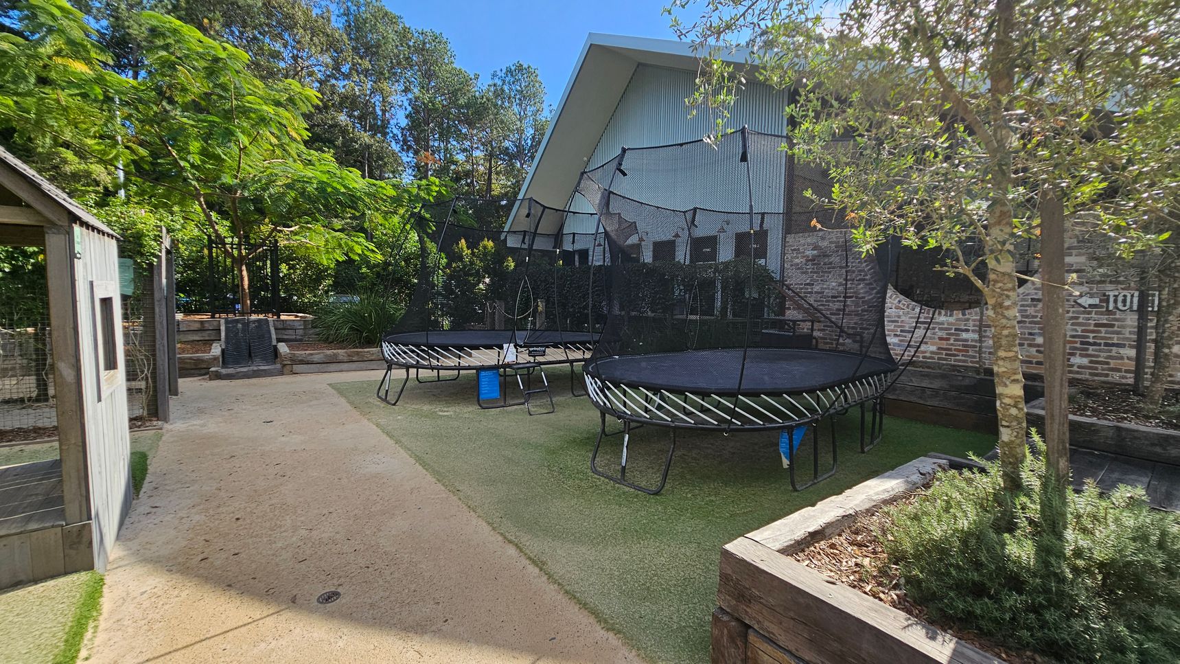 A garden area with two trampolines in the centre, each surrounded by safety nets. On the left, there's a small wooden structure resembling a shed. The ground is a mix of concrete and artificial grass. In the foreground, a planter box with a small tree and shrubs. The background features a large building with a slanted roof and brick walls, surrounded by tall trees and greenery. A garden area with two trampolines in the centre, each surrounded by safety nets. On the left, there's a small wooden structure resembling a shed. The ground is a mix of concrete and artificial grass. In the foreground, a planter box with a small tree and shrubs. The background features a large building with a slanted roof and brick walls, surrounded by tall trees and greenery.