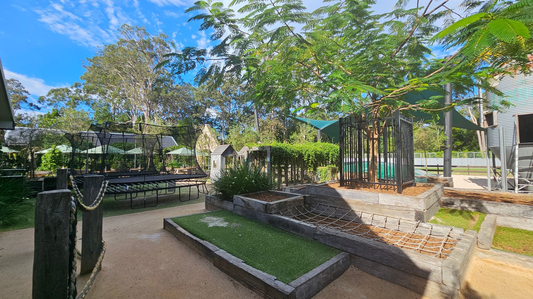 Outdoor play area with various structures. On the left, a large trampoline with a netted enclosure is surrounded by wooden posts connected by ropes. In the centre, a small wooden playhouse and a garden with lush greenery. On the right, a tree is enclosed by a metal fence, and a climbing net is attached to raised wooden platforms. The ground is a mix of sand and artificial grass. In the background, tall trees and green shade sails provide cover. Outdoor play area with various structures. On the left, a large trampoline with a netted enclosure is surrounded by wooden posts connected by ropes. In the centre, a small wooden playhouse and a garden with lush greenery. On the right, a tree is enclosed by a metal fence, and a climbing net is attached to raised wooden platforms. The ground is a mix of sand and artificial grass. In the background, tall trees and green shade sails provide cover.