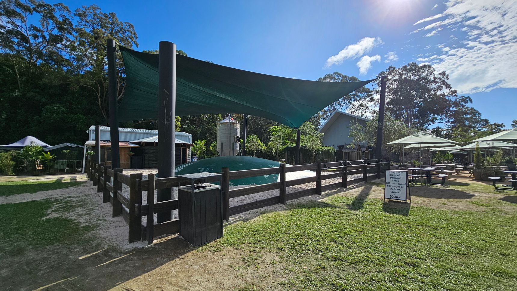 A fenced area with a large, inflated play structure covered by a green shade sail is in the centre. To the left, there are small buildings with metal roofs surrounded by trees. On the right, outdoor seating with tables and umbrellas is visible on a grassy area. A sign with text is placed near the fence. The background features tall trees and a clear sky. The ground is a mix of grass and dirt. A fenced area with a large, inflated play structure covered by a green shade sail is in the centre. To the left, there are small buildings with metal roofs surrounded by trees. On the right, outdoor seating with tables and umbrellas is visible on a grassy area. A sign with text is placed near the fence. The background features tall trees and a clear sky. The ground is a mix of grass and dirt.