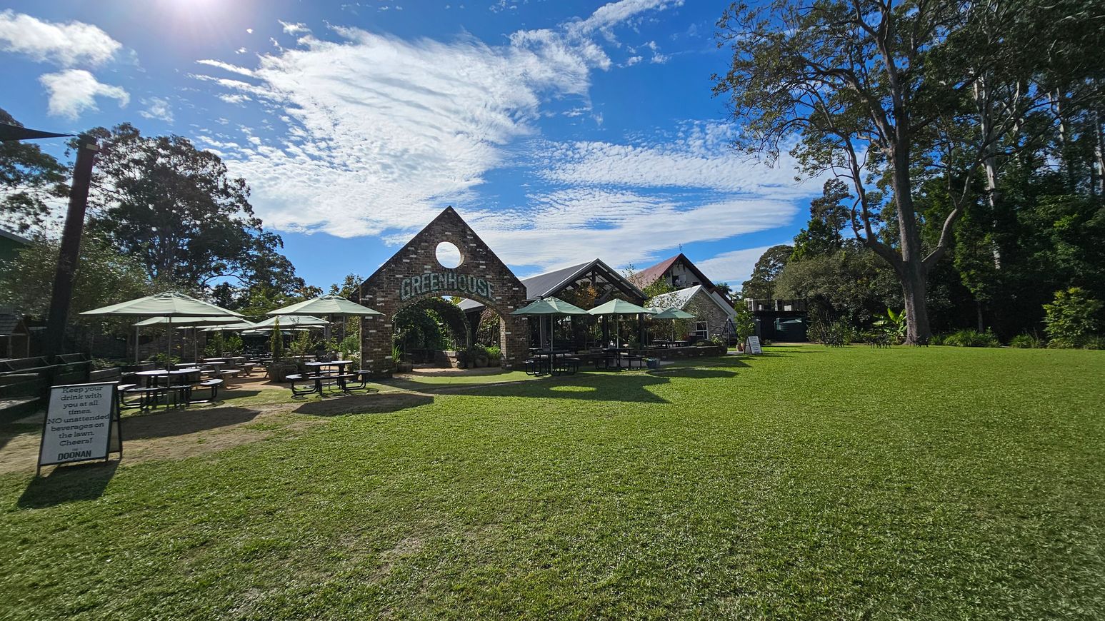 A grassy area with a building in the centre featuring a triangular roof and a circular window, labelled "GREENHOUSE". Several outdoor tables with umbrellas are on the left and right. A sign on the left reads, "Keep your drink with you at all times. NO unattended beverages on the lawn. Cheers! DOONAN". Tall trees surround the area, providing shade and greenery. A grassy area with a building in the centre featuring a triangular roof and a circular window, labelled "GREENHOUSE". Several outdoor tables with umbrellas are on the left and right. A sign on the left reads, "Keep your drink with you at all times. NO unattended beverages on the lawn. Cheers! DOONAN". Tall trees surround the area, providing shade and greenery.