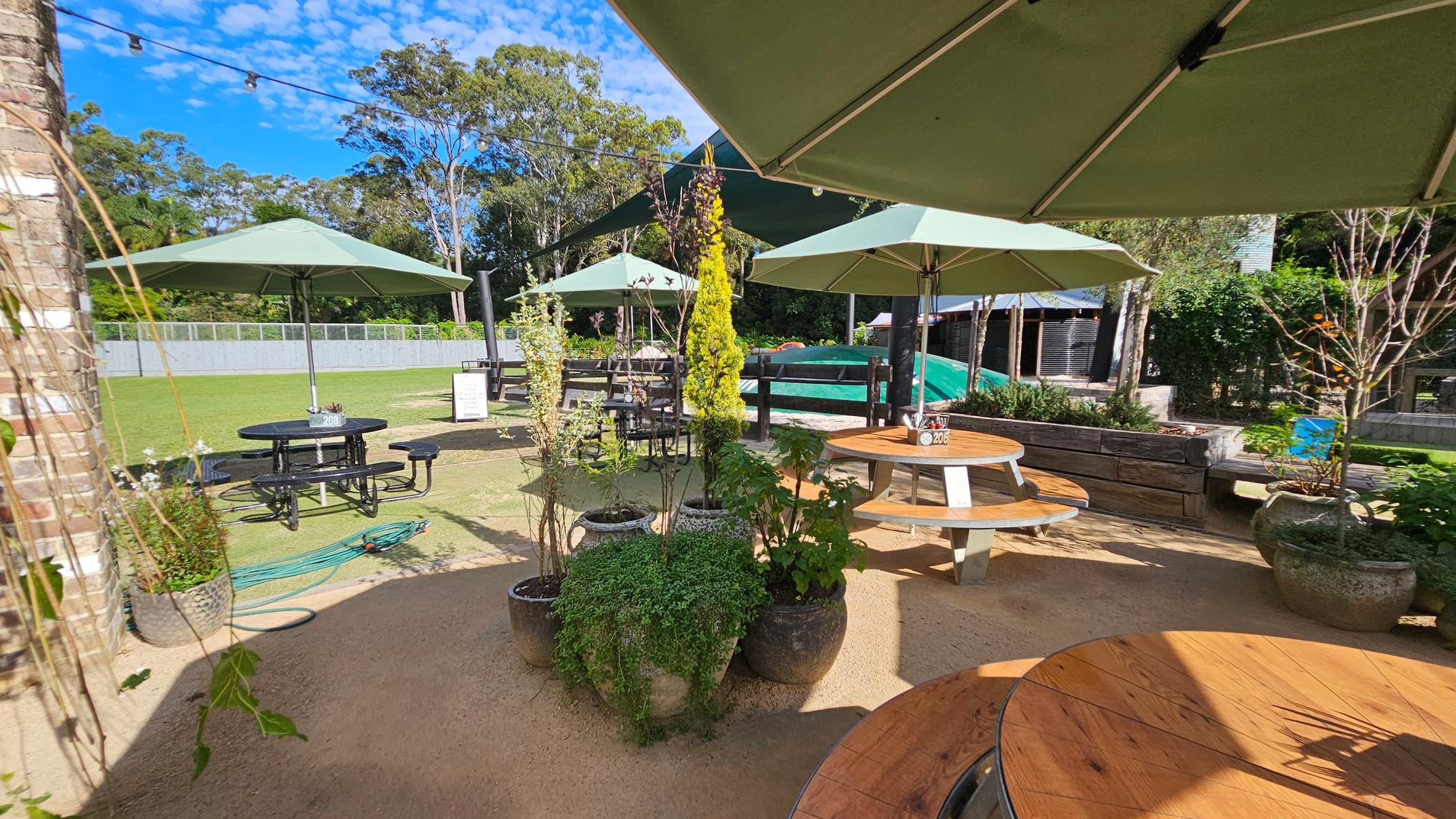 Outdoor seating area with round wooden tables and green umbrellas. In the foreground, several potted plants are arranged on a sandy surface. To the left, a brick wall is partially visible. In the background, a grassy area with more tables and a fence is surrounded by tall trees. A garden hose is coiled on the ground near a black metal table. Overhead, string lights are strung across the area. Outdoor seating area with round wooden tables and green umbrellas. In the foreground, several potted plants are arranged on a sandy surface. To the left, a brick wall is partially visible. In the background, a grassy area with more tables and a fence is surrounded by tall trees. A garden hose is coiled on the ground near a black metal table. Overhead, string lights are strung across the area.