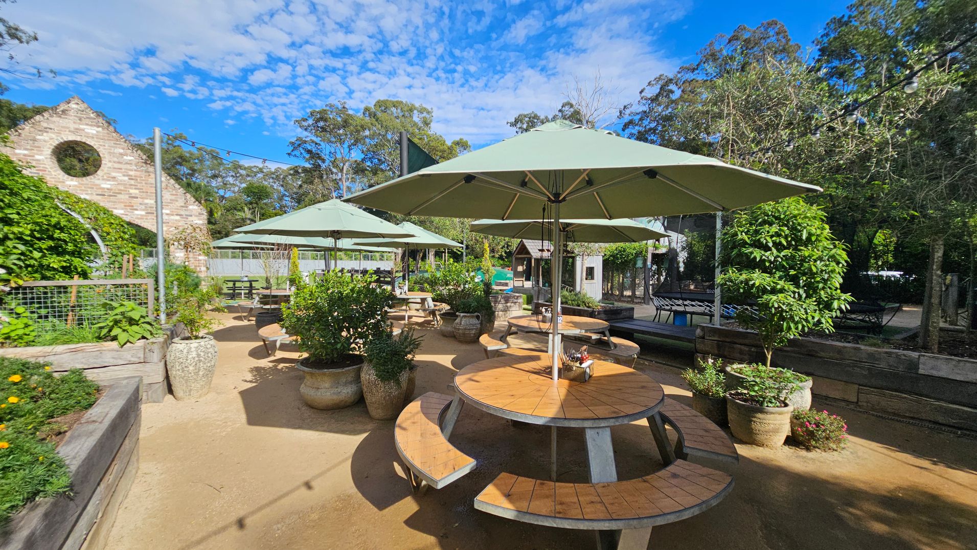 Outdoor seating area with round wooden tables and green umbrellas in the centre. Potted plants and greenery surround the space. On the left, a brick structure with a circular window is partially covered in vines. In the background, trees and a wooden fence are visible. The ground is sandy, and string lights are strung overhead. Outdoor seating area with round wooden tables and green umbrellas in the centre. Potted plants and greenery surround the space. On the left, a brick structure with a circular window is partially covered in vines. In the background, trees and a wooden fence are visible. The ground is sandy, and string lights are strung overhead.