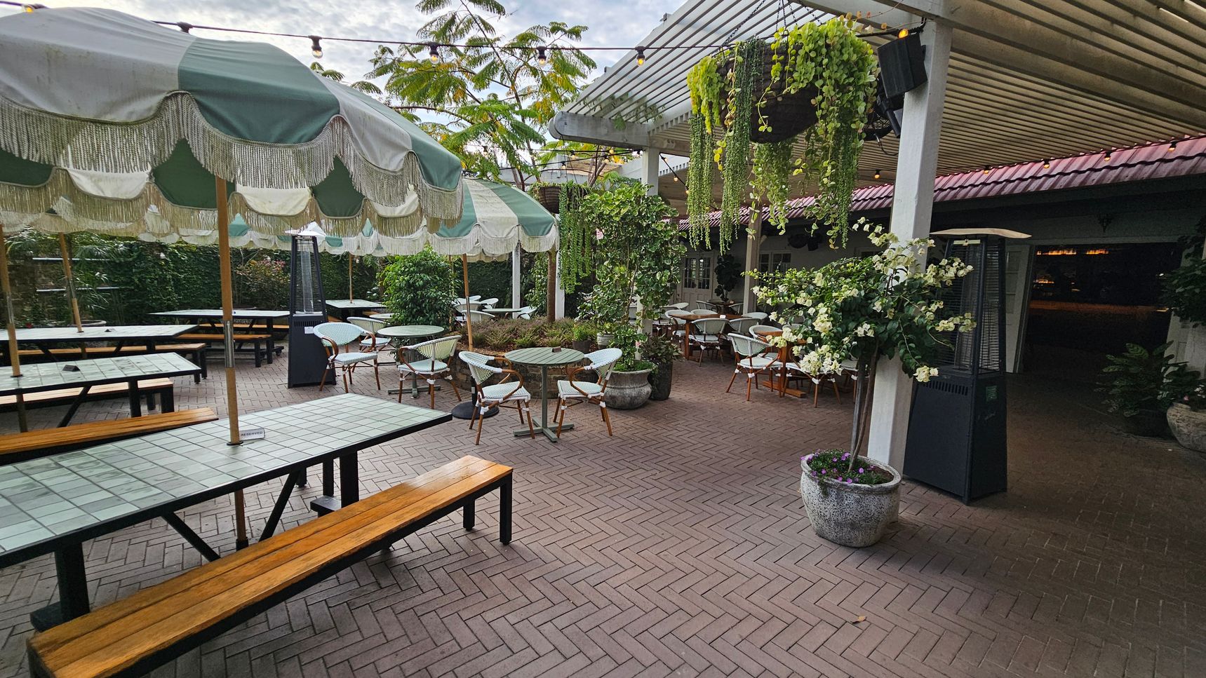 Outdoor seating area with a herringbone-patterned brick floor. On the left, there are long tables with wooden benches under large green and white striped umbrellas. In the centre, round tables with wicker chairs are surrounded by potted plants. On the right, a pergola with hanging plants provides shade. A tall outdoor heater stands nearby. The background features lush greenery and a building with a red-tiled roof. Outdoor seating area with a herringbone-patterned brick floor. On the left, there are long tables with wooden benches under large green and white striped umbrellas. In the centre, round tables with wicker chairs are surrounded by potted plants. On the right, a pergola with hanging plants provides shade. A tall outdoor heater stands nearby. The background features lush greenery and a building with a red-tiled roof.
