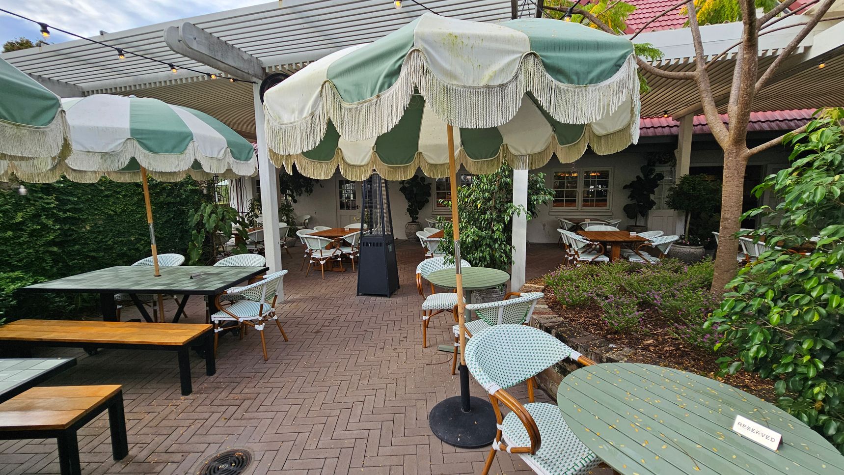 Outdoor café setting with green and white striped umbrellas featuring fringe, covering round tables with wicker chairs. A "Reserved" sign is on one table. Wooden benches accompany rectangular tables on the left. The ground is paved with herringbone-patterned bricks. A pergola with string lights is overhead. Lush greenery and potted plants surround the area, with a tree on the right. A building with a red-tiled roof and windows is in the background. Outdoor café setting with green and white striped umbrellas featuring fringe, covering round tables with wicker chairs. A "Reserved" sign is on one table. Wooden benches accompany rectangular tables on the left. The ground is paved with herringbone-patterned bricks. A pergola with string lights is overhead. Lush greenery and potted plants surround the area, with a tree on the right. A building with a red-tiled roof and windows is in the background.