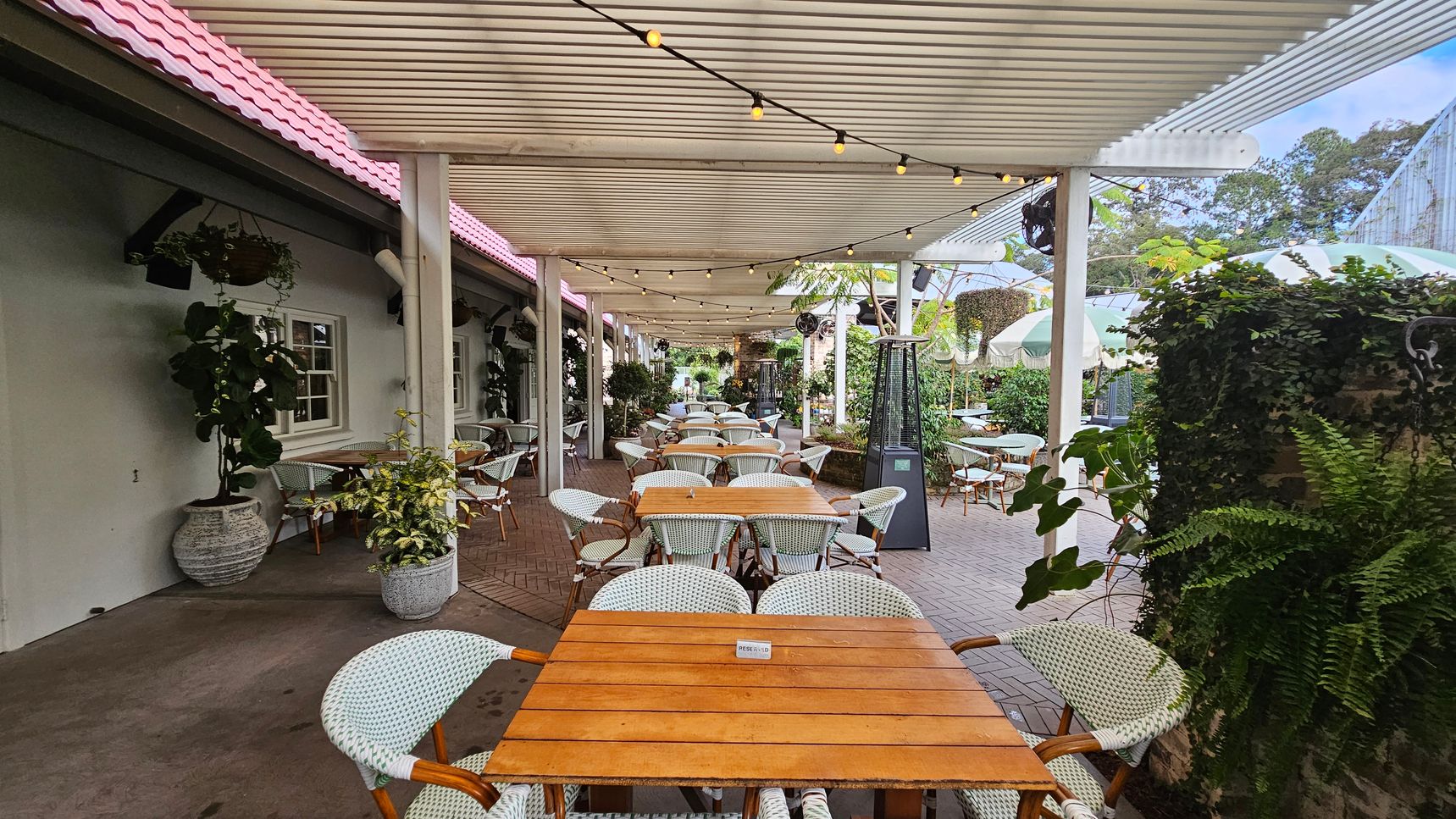 Outdoor dining area with wooden tables and white wicker chairs arranged in rows. A pergola with string lights covers the space. On the left, a building with a pink-tiled roof and green walls features windows and hanging plants. On the right, green foliage and potted plants line the area. The floor is a mix of concrete and brick, and patio heaters are visible. In the background, more tables and greenery are present, creating a lush atmosphere. Outdoor dining area with wooden tables and white wicker chairs arranged in rows. A pergola with string lights covers the space. On the left, a building with a pink-tiled roof and green walls features windows and hanging plants. On the right, green foliage and potted plants line the area. The floor is a mix of concrete and brick, and patio heaters are visible. In the background, more tables and greenery are present, creating a lush atmosphere.