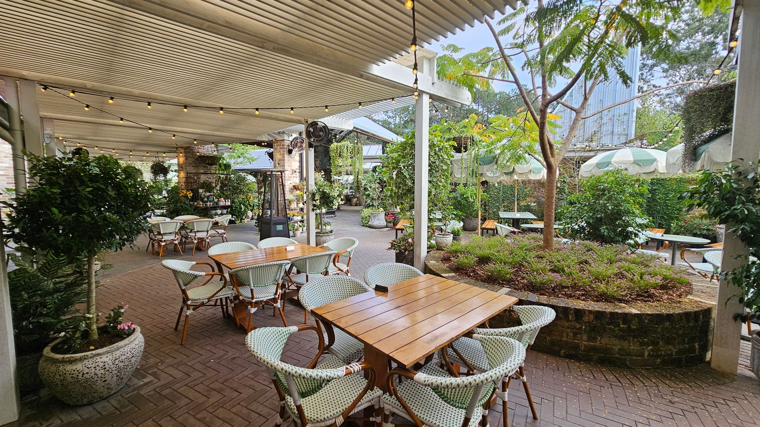 An outdoor dining area with wooden tables and wicker chairs on a herringbone brick floor. The left side features a covered patio with string lights and potted plants. In the centre, a round brick planter with a small tree and shrubs. The right side has more tables under green and white striped umbrellas. The background includes lush greenery and a tall building partially visible. The ceiling is a slatted pergola. An outdoor dining area with wooden tables and wicker chairs on a herringbone brick floor. The left side features a covered patio with string lights and potted plants. In the centre, a round brick planter with a small tree and shrubs. The right side has more tables under green and white striped umbrellas. The background includes lush greenery and a tall building partially visible. The ceiling is a slatted pergola.