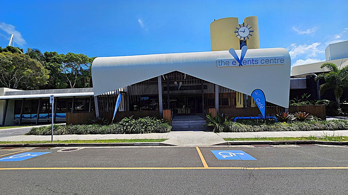 A modern building with a curved white roof and a sign reading "the events centre" on the front. A clock is mounted on a yellow cylindrical structure above. Blue flags with "café" are displayed near the entrance. The foreground features a road with marked disabled parking spaces and a pedestrian path lined with greenery. Trees are visible to the left, and palm trees are on the right. A modern building with a curved white roof and a sign reading "the events centre" on the front. A clock is mounted on a yellow cylindrical structure above. Blue flags with "café" are displayed near the entrance. The foreground features a road with marked disabled parking spaces and a pedestrian path lined with greenery. Trees are visible to the left, and palm trees are on the right.