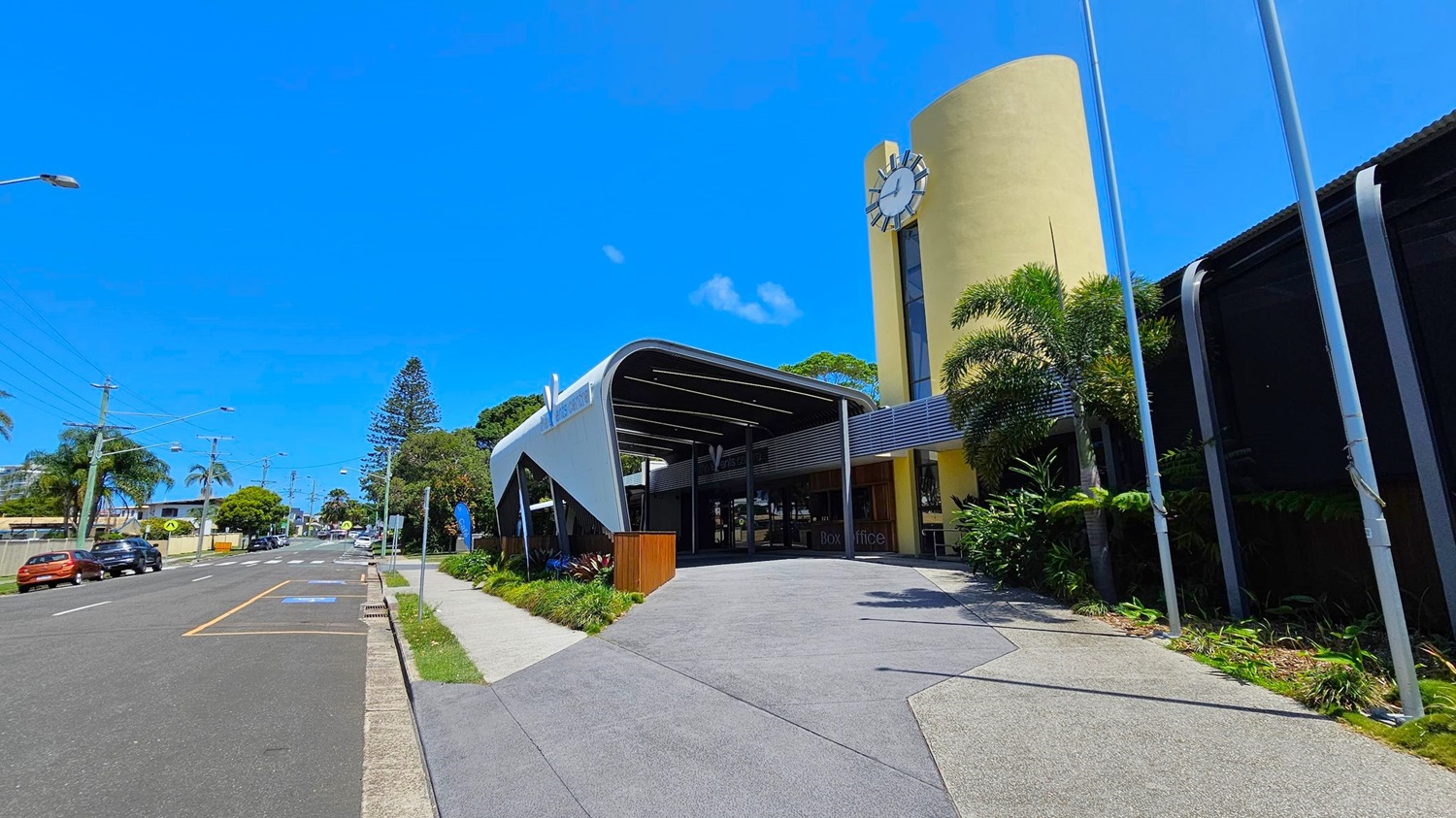 A modern building with a curved entrance and a large clock on a yellow cylindrical tower on the right. The foreground features a wide pavement with greenery along the edge. On the left, a street lined with parked cars and palm trees extends into the distance. The building has a sleek, contemporary design with a mix of metal and glass elements. A modern building with a curved entrance and a large clock on a yellow cylindrical tower on the right. The foreground features a wide pavement with greenery along the edge. On the left, a street lined with parked cars and palm trees extends into the distance. The building has a sleek, contemporary design with a mix of metal and glass elements.