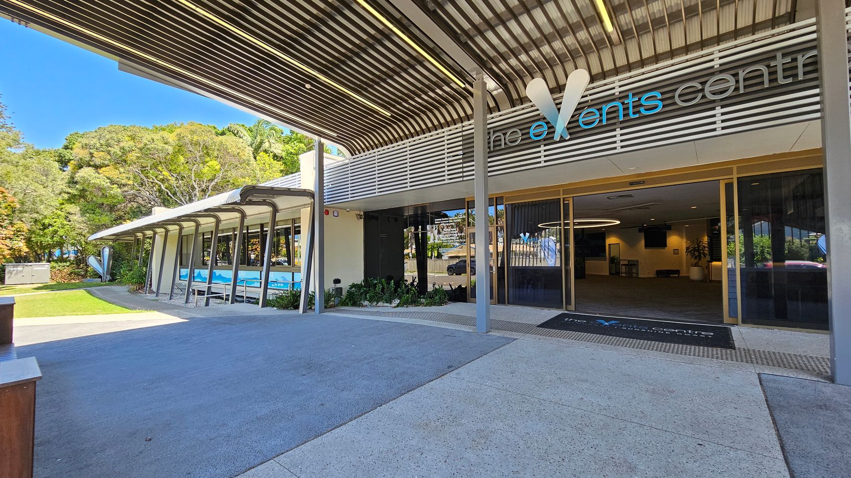 Entrance to a modern events centre with a large overhanging roof and glass doors. The sign above reads "the events centre" with stylised lettering. On the left, a pathway leads past a building with large windows and a mural, surrounded by lush greenery and trees. The ground is paved with light-coloured concrete, and the ceiling features a metal slatted design. Inside, a circular light fixture is visible. Entrance to a modern events centre with a large overhanging roof and glass doors. The sign above reads "the events centre" with stylised lettering. On the left, a pathway leads past a building with large windows and a mural, surrounded by lush greenery and trees. The ground is paved with light-coloured concrete, and the ceiling features a metal slatted design. Inside, a circular light fixture is visible.